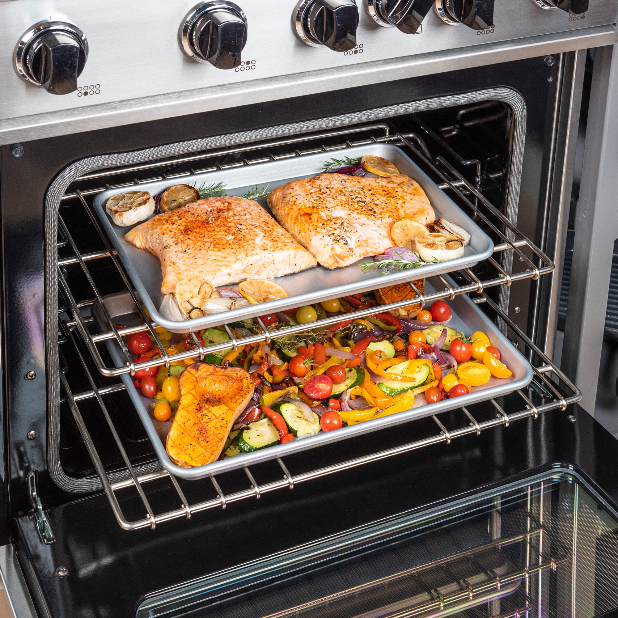 In the oven, two trays from the Viking 2-Piece Nonstick Aluminized Steel Baking Sheet Set by Viking Culinary Products hold seasoned salmon with veggies on top and a vibrant medley of roasted tomatoes, squash, and peppers below.