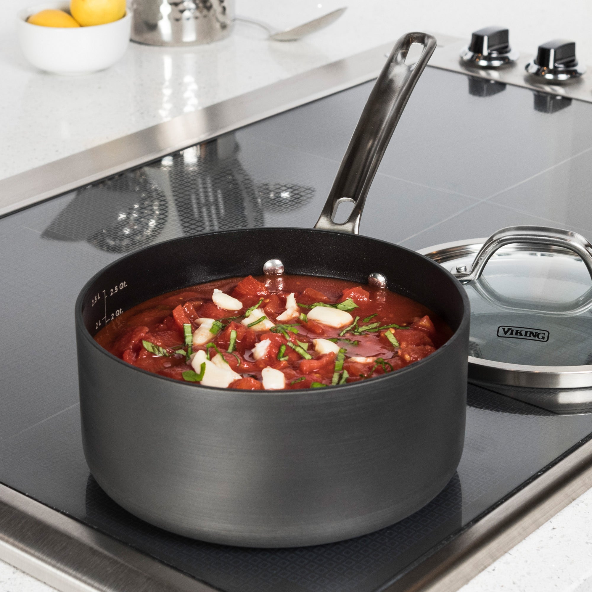 A Viking Hard Anodized Nonstick 3-Quart Sauce Pan by Viking Culinary Products, filled with tomato stew and garnished with herbs and garlic, sits on a modern stovetop. The glass lid is nearby, with lemons visible in the background.