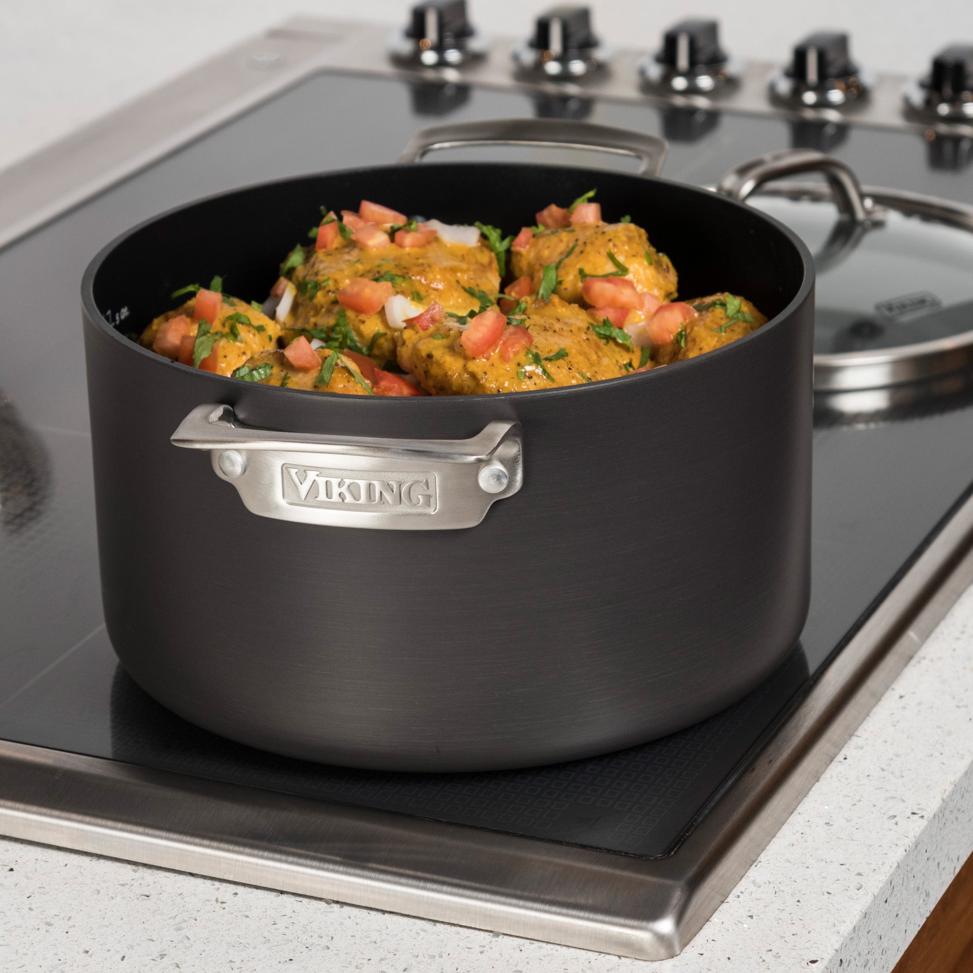 The Viking Culinary Products Hard Anodized Nonstick 6-Quart Dutch Oven with Glass Lid sits on a stovetop, filled with cooked food like meat or dumplings topped with chopped tomatoes and herbs.