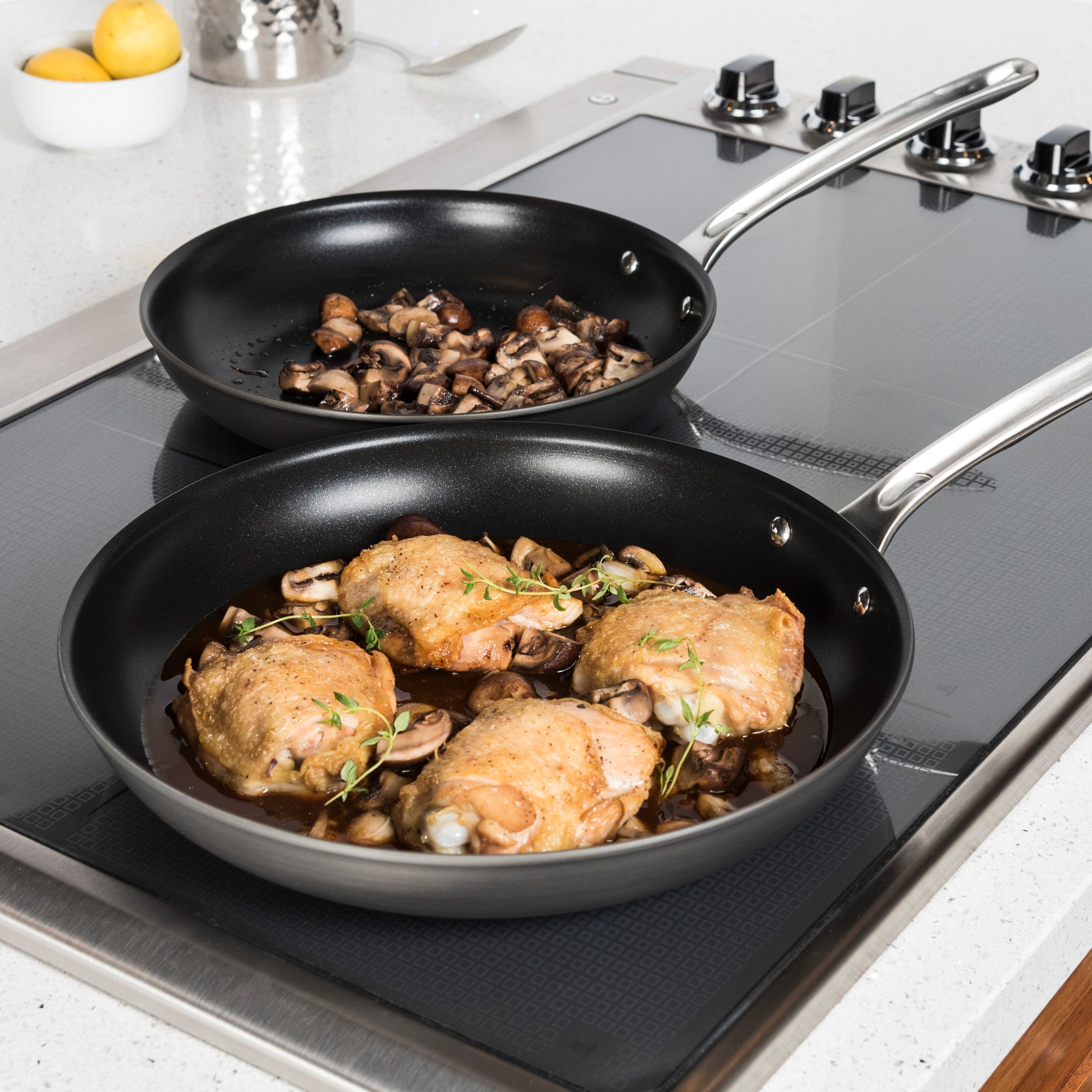 The Viking Hard Anodized Nonstick 2-Piece Fry Pan Set by Viking Culinary Products shines on the stovetop, one pan filled with chicken thighs, mushrooms, and herbs, the other with sautéed mushrooms. Both feature signature Stay-Cool Handles. Lemons and a metal container sit nearby.