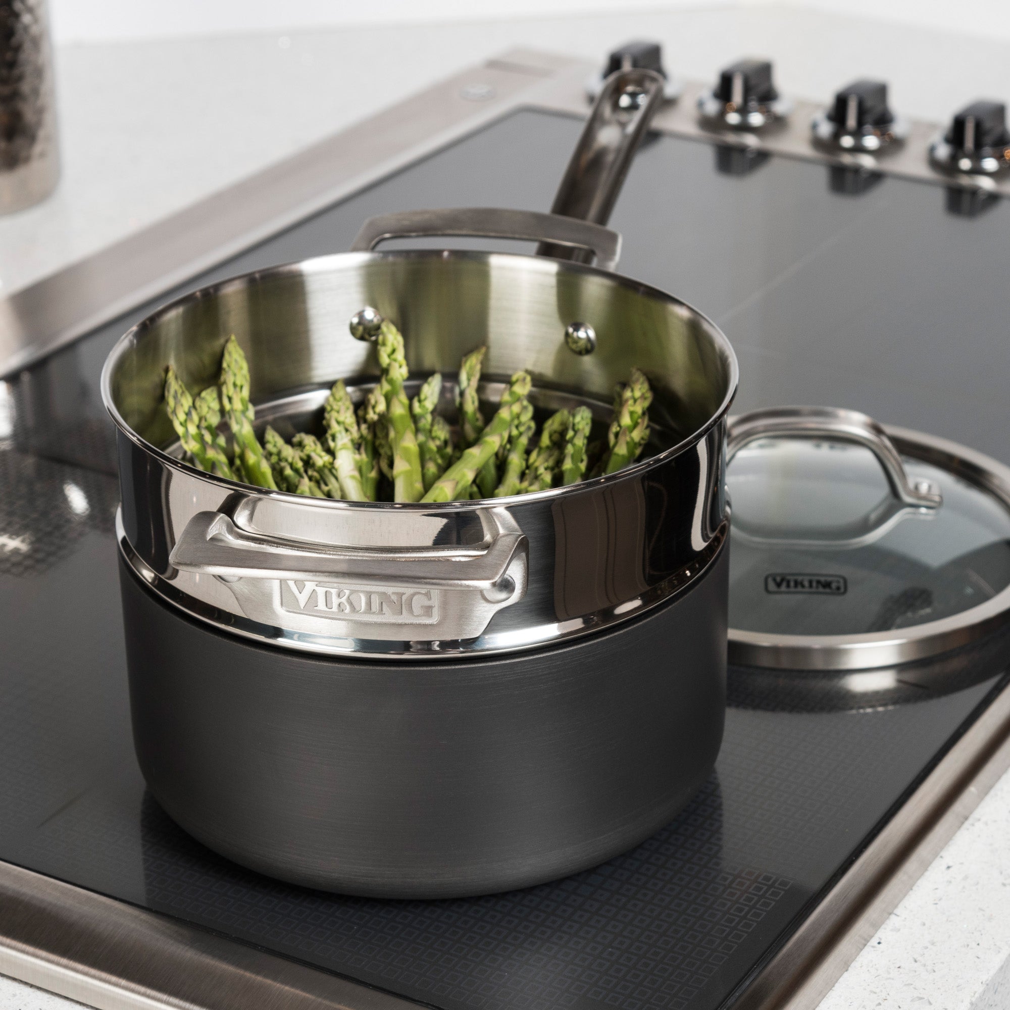 Fresh asparagus spears stand upright in the Viking Stainless Steel Universal Steamer Insert and Colander by Viking Culinary Products, with its glass lid set aside on a modern stovetop.