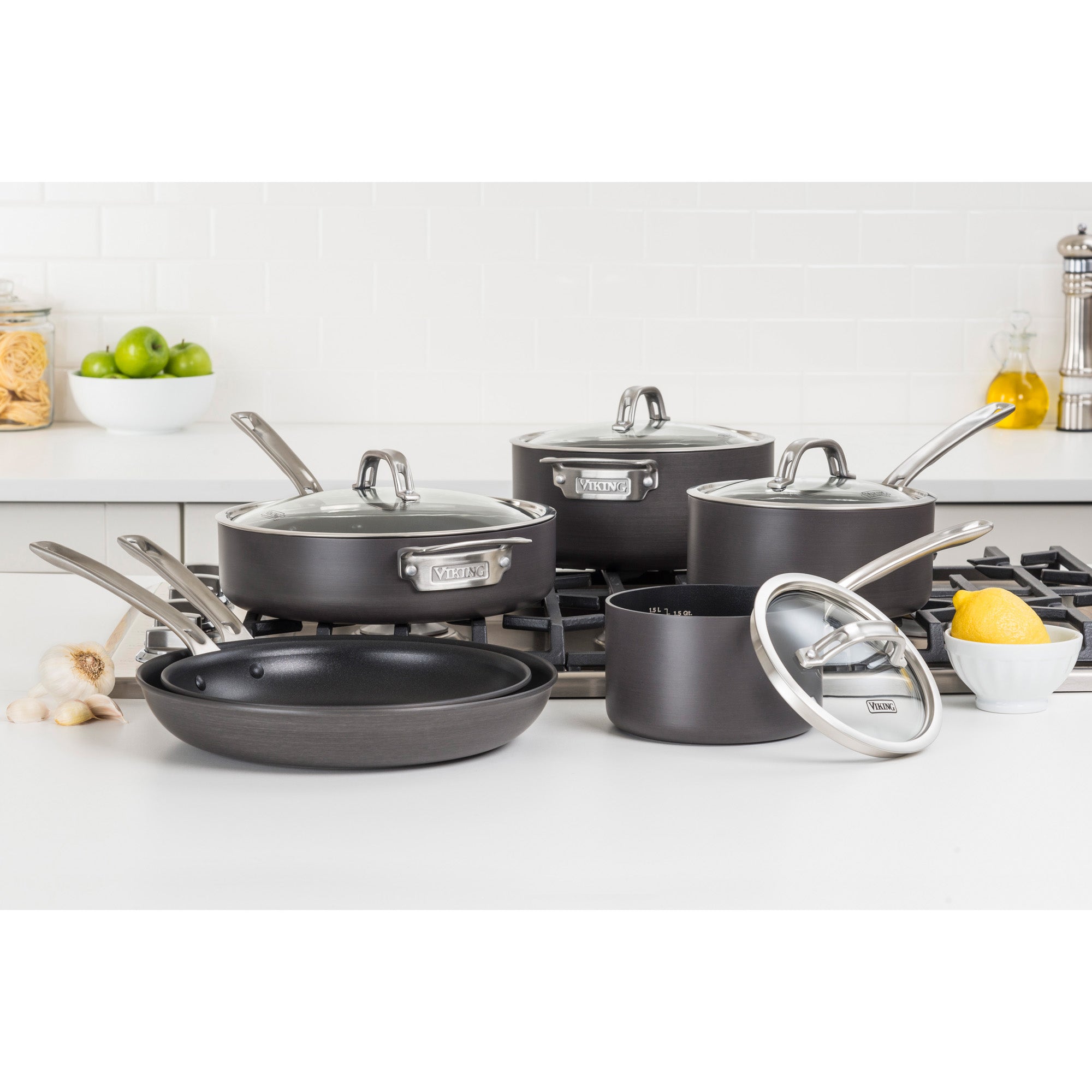The Viking Hard Anodized Nonstick 10-Piece Cookware Set with Glass Lids by Viking Culinary Products is displayed on a modern kitchen counter and stove, surrounded by lemons, apples, garlic, and pasta.