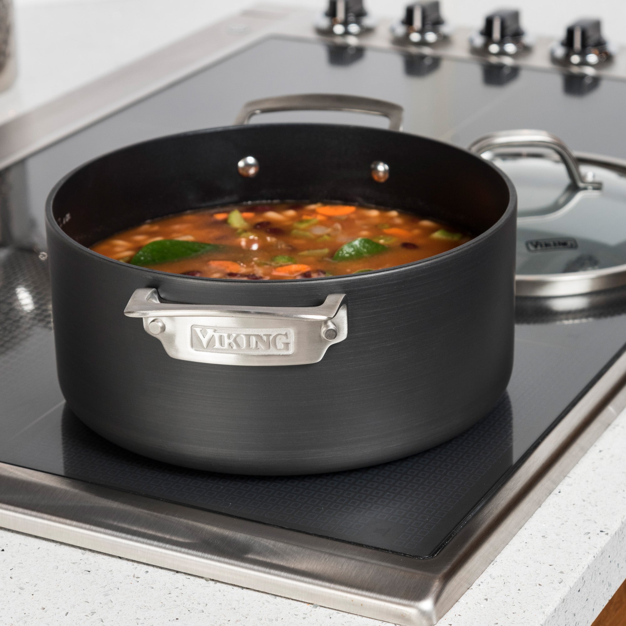 A Viking Culinary Products Hard Anodized Nonstick Dutch Oven from the 10-Piece Cookware Set holds vegetable soup with lime slices on a modern stovetop, with the glass lid placed nearby.