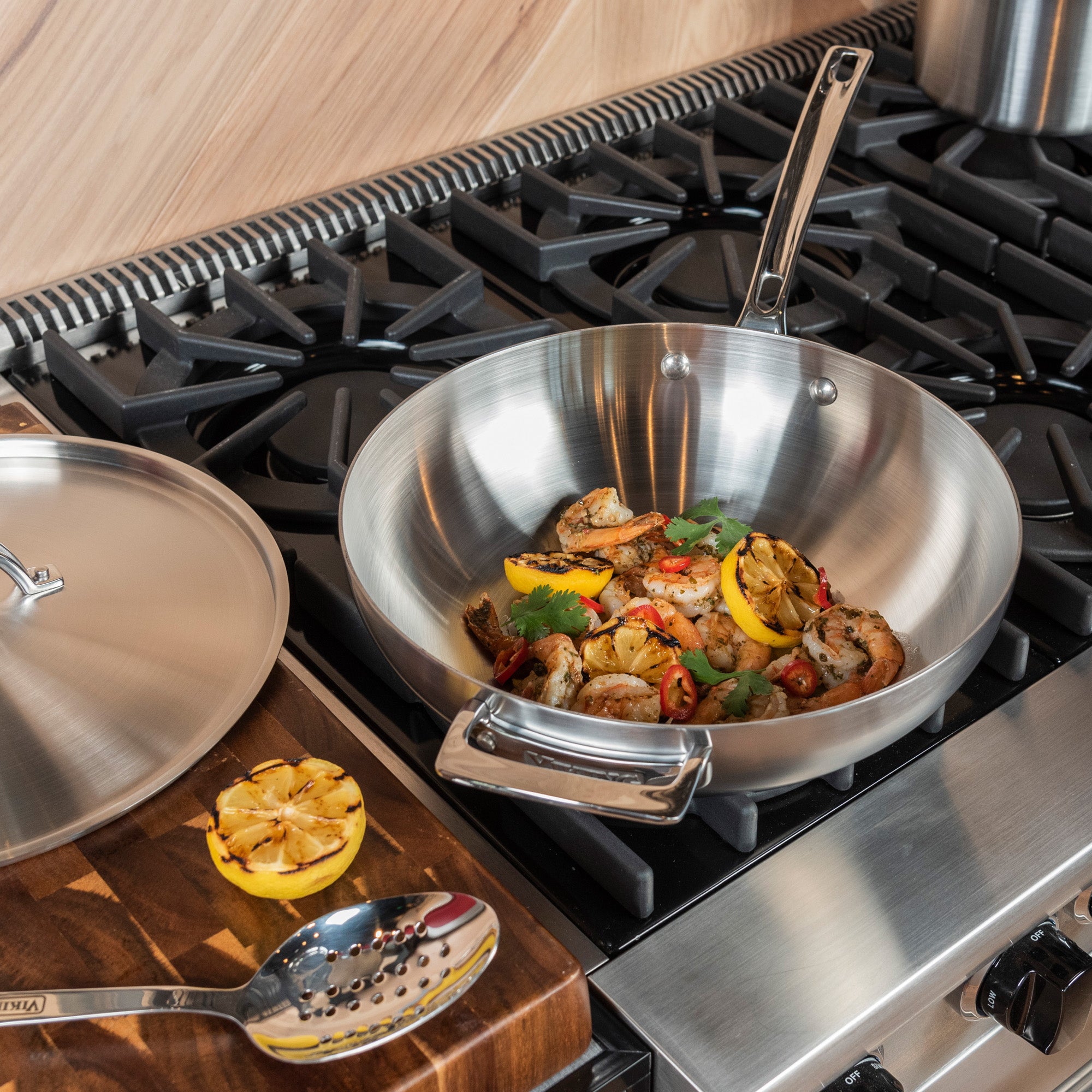 The Viking Professional 5-Ply 3-Quart Saucier with Metal Lid by Viking Culinary Products holds cooked shrimp, lemon, and herbs on a gas stove; its lid, a slotted spoon, and grilled lemon are on a nearby wooden cutting board.