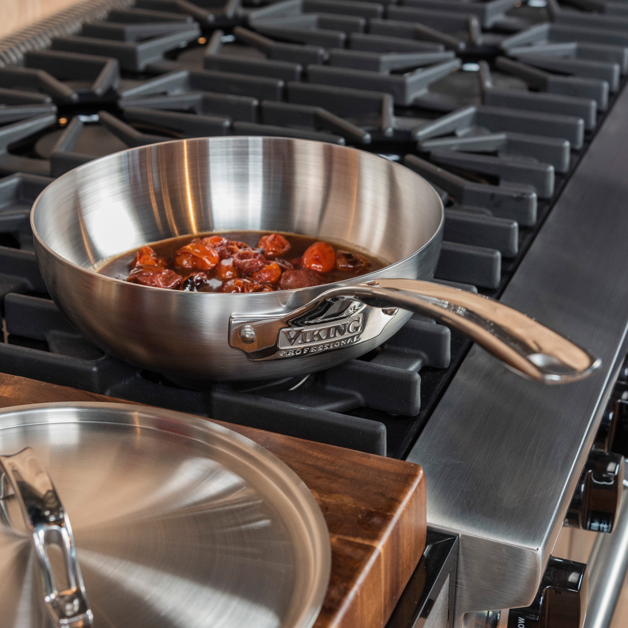 A Viking Culinary Products Professional 5-Ply Stainless Steel 12-Inch/5.6-Quart Chefs Pan with Metal Lid simmers tomato sauce on a gas stove, while the matching lid rests on a wooden surface nearby.