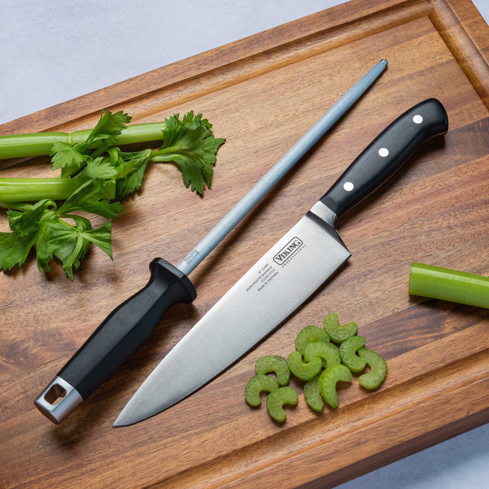 A Viking Professional 8-Inch Chefs Knife by Viking Culinary Products and a honing steel are displayed on a wooden cutting board with fresh celery stalks and slices nearby.