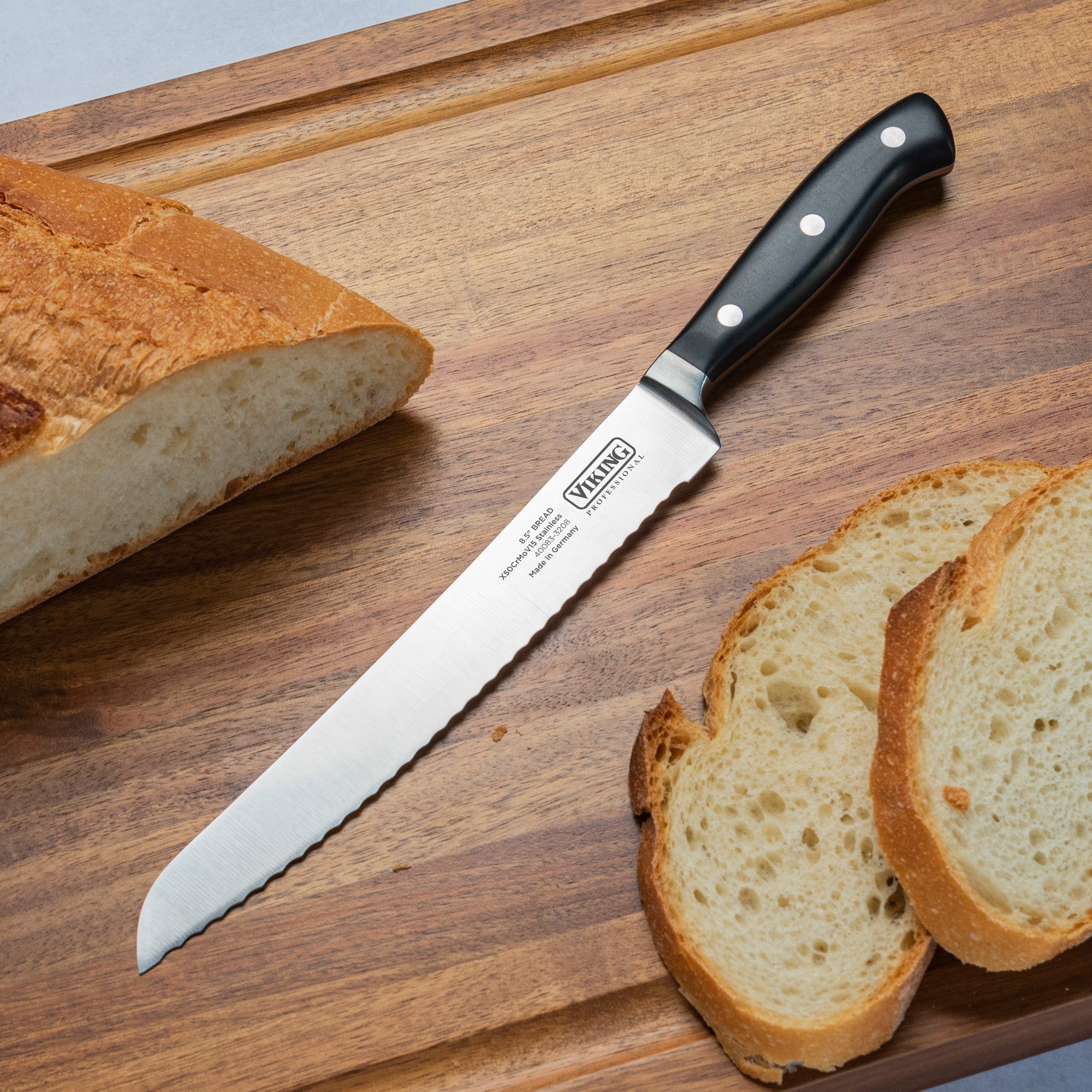 The Viking Professional 8-Inch Bread Knife by Viking Culinary Products, featuring a black handle and German steel blade, rests on a wooden cutting board beside a partially sliced loaf of bread and two slices.