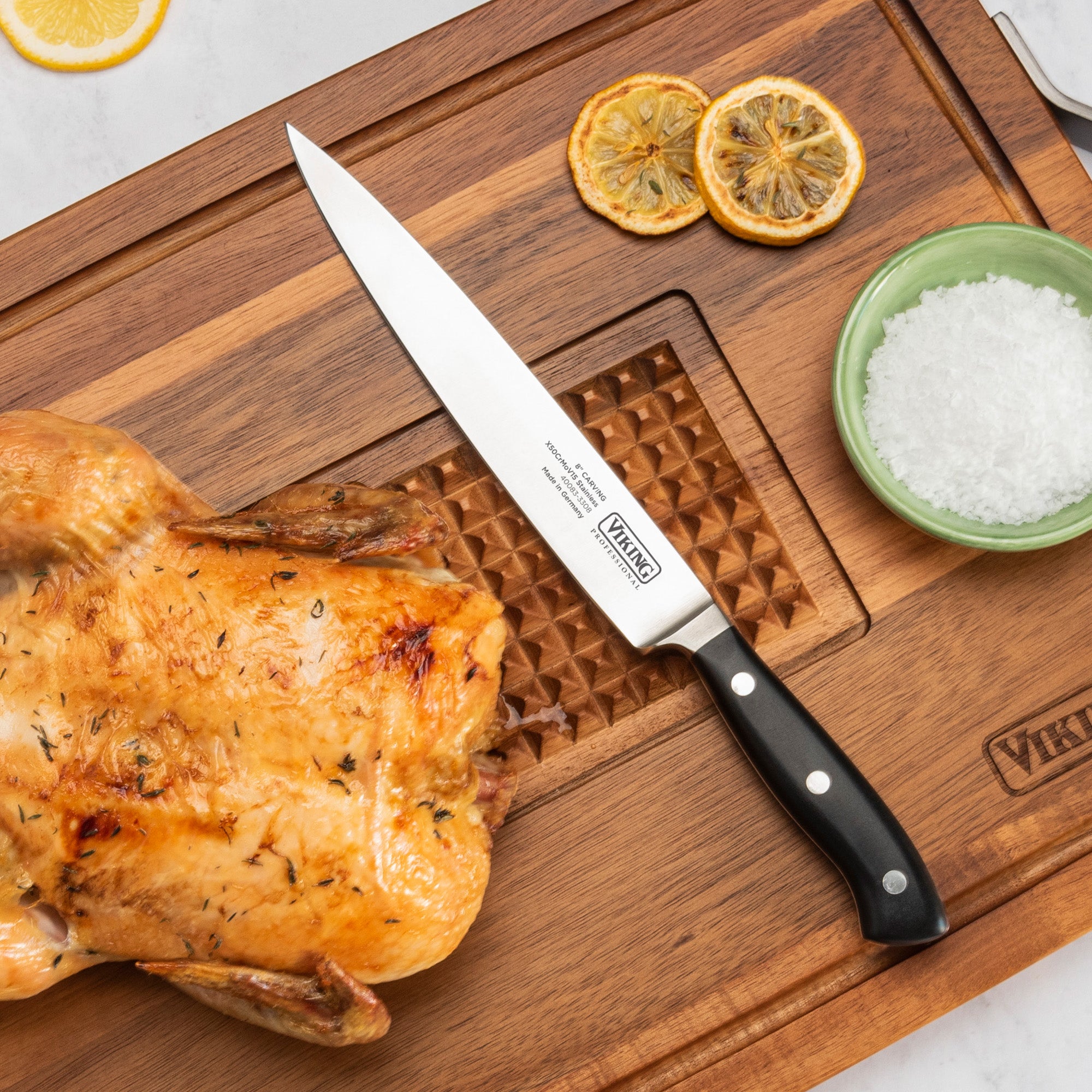 A whole roasted chicken rests on a wooden cutting board beside a Viking Professional 8 Carving Knife from Viking Culinary Products, a green bowl of coarse salt, and two slices of dried lemon.