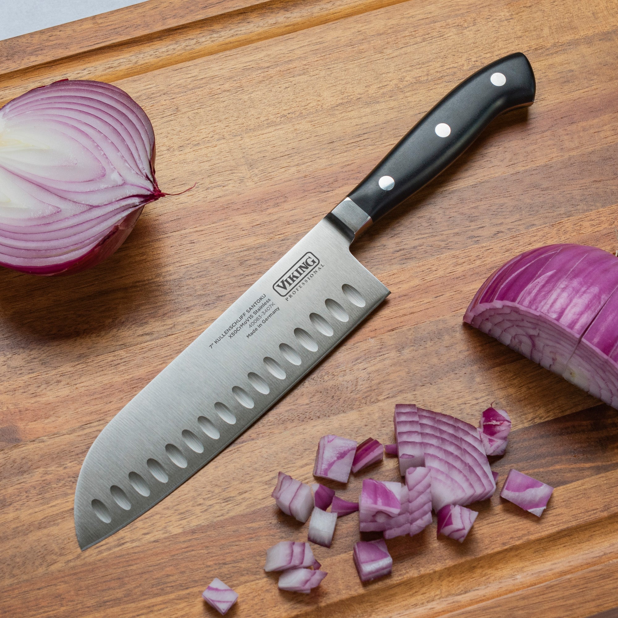 The Viking Professional 7-Inch Santoku Knife by Viking Culinary Products, featuring a black handle and German steel blade, rests on a wooden cutting board beside chopped, sliced, and halved pieces of red onion.