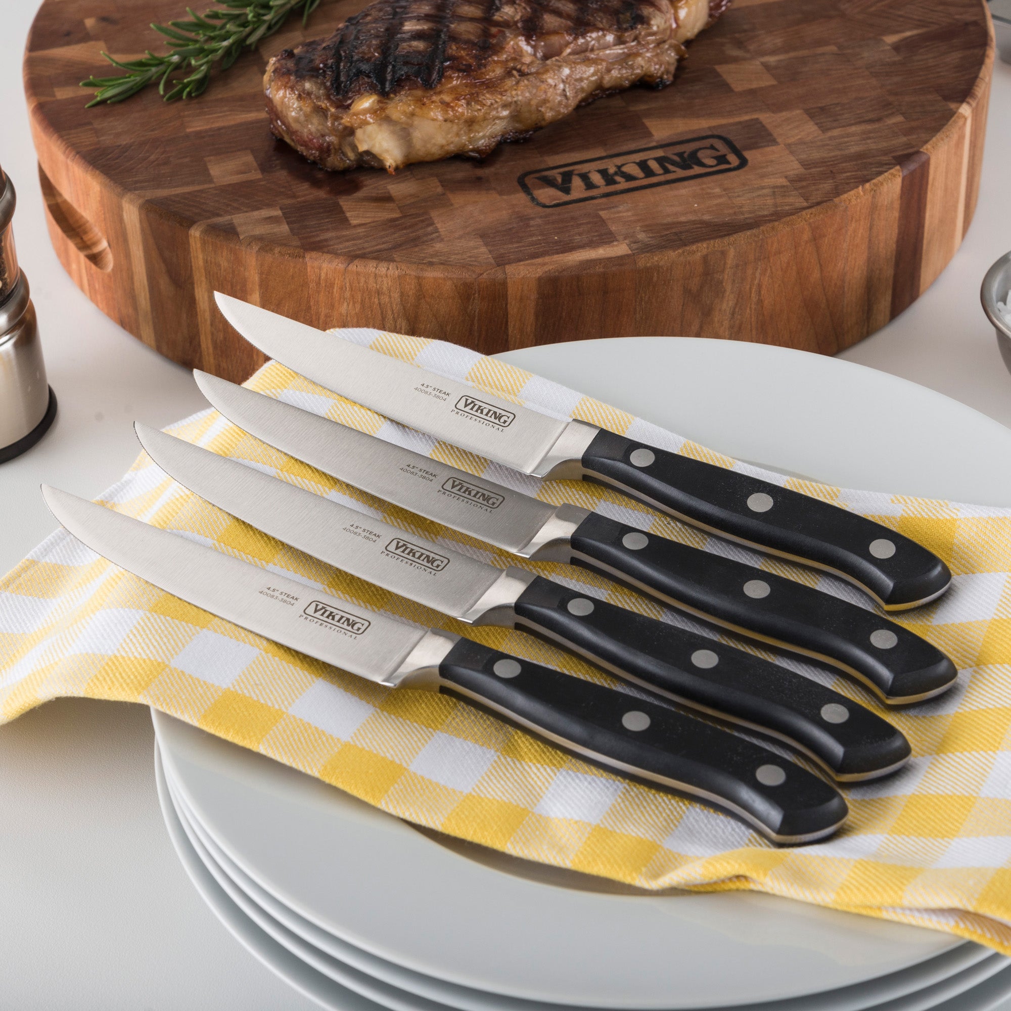 The Viking Professional 4-Piece Steak Knife Set by Viking Culinary Products, featuring black handles and a full-tang design, rests on a yellow and white checkered cloth with a stack of plates; a grilled steak sits on a wooden cutting board in the background.