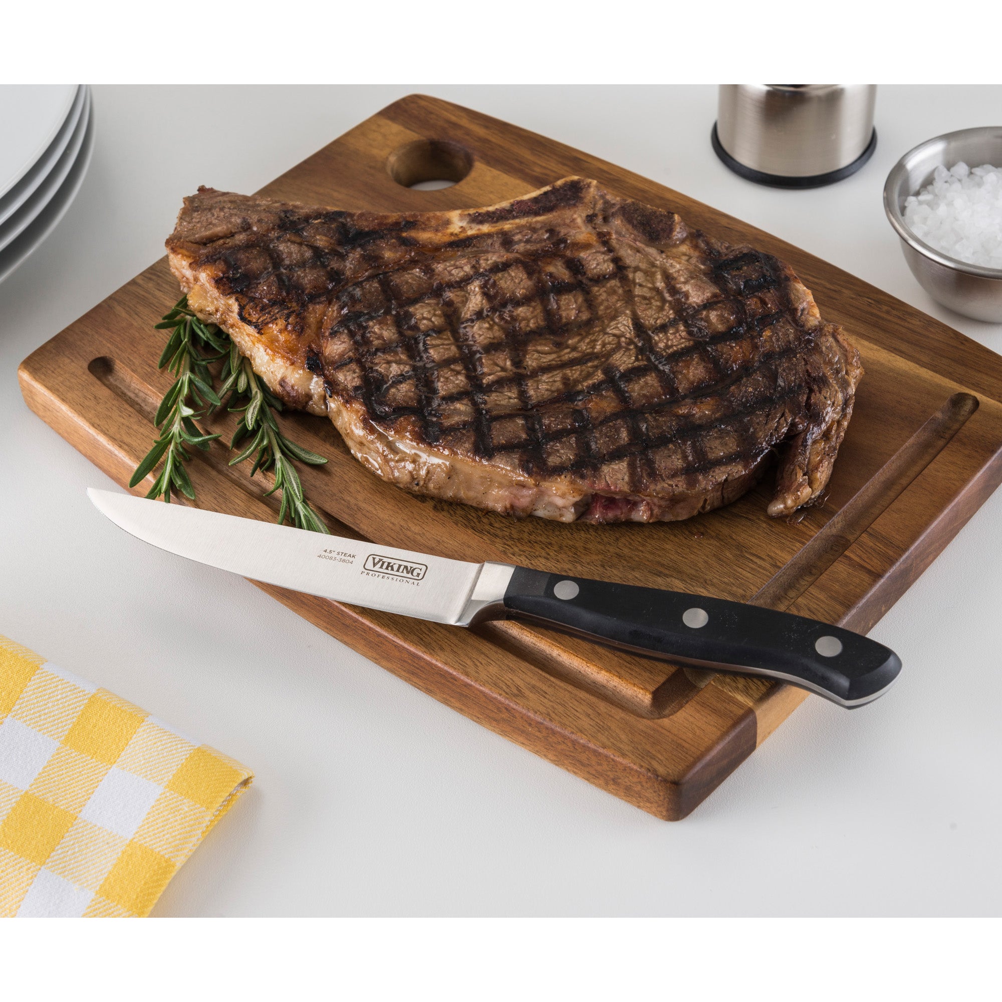A grilled steak with grill marks sits on a wooden cutting board, garnished with rosemary. A Viking Professional 4.5-Inch Steak Knife by Viking Culinary Products rests beside it. Plates, salt, and a yellow checkered napkin are nearby on the white table.