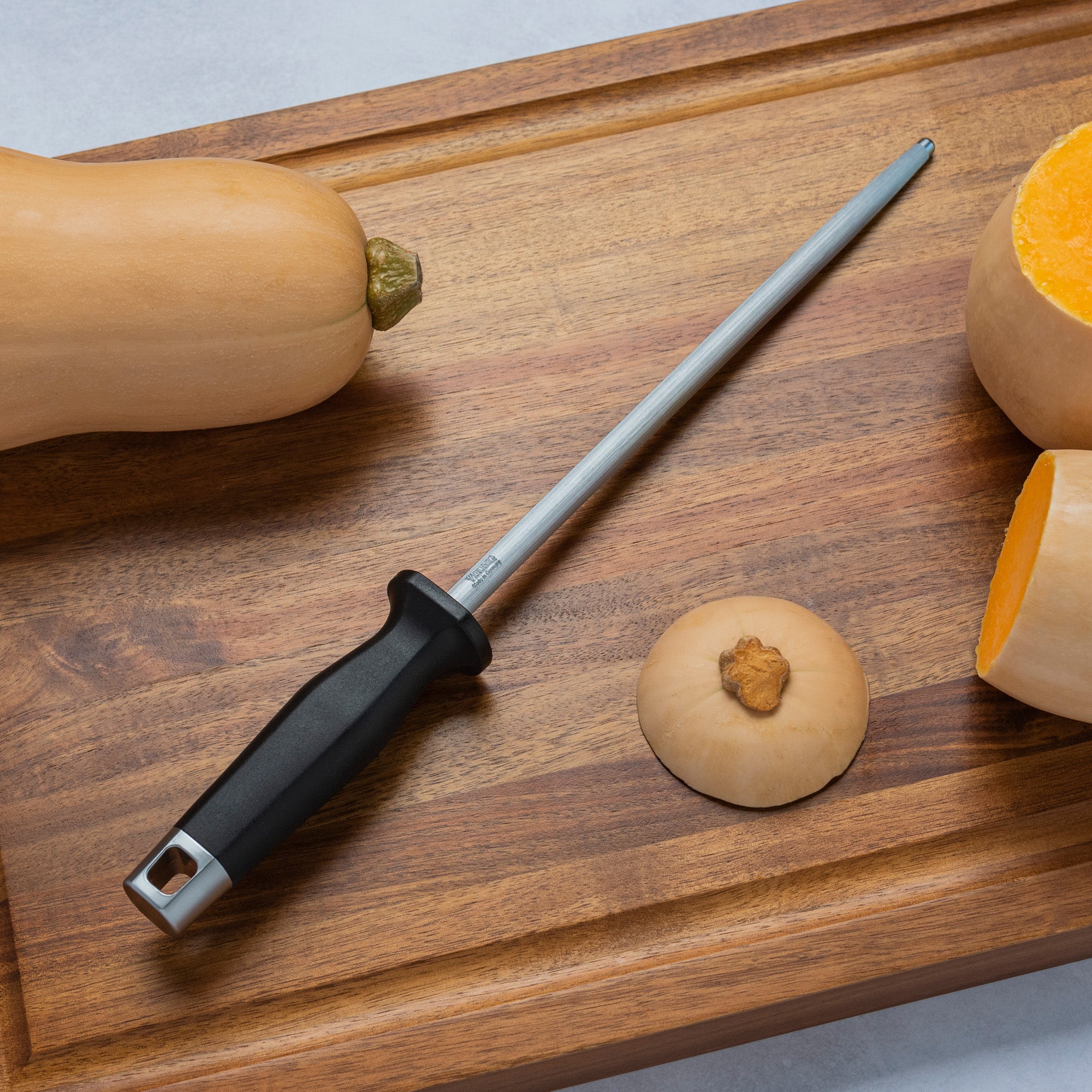 The Viking Professional 10-Inch Sharpening Steel by Viking Culinary Products, with a durable black handle, lies on a wooden cutting board next to a whole butternut squash, sliced squash pieces, and the top.