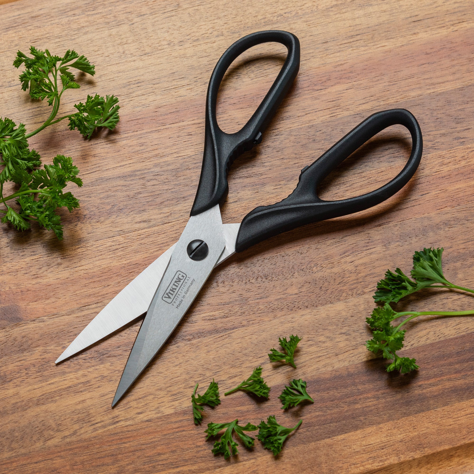 A pair of Viking Professional 8-Inch Scissors by Viking Culinary Products, with black handles, lies open on a wooden surface surrounded by fresh parsley sprigs.