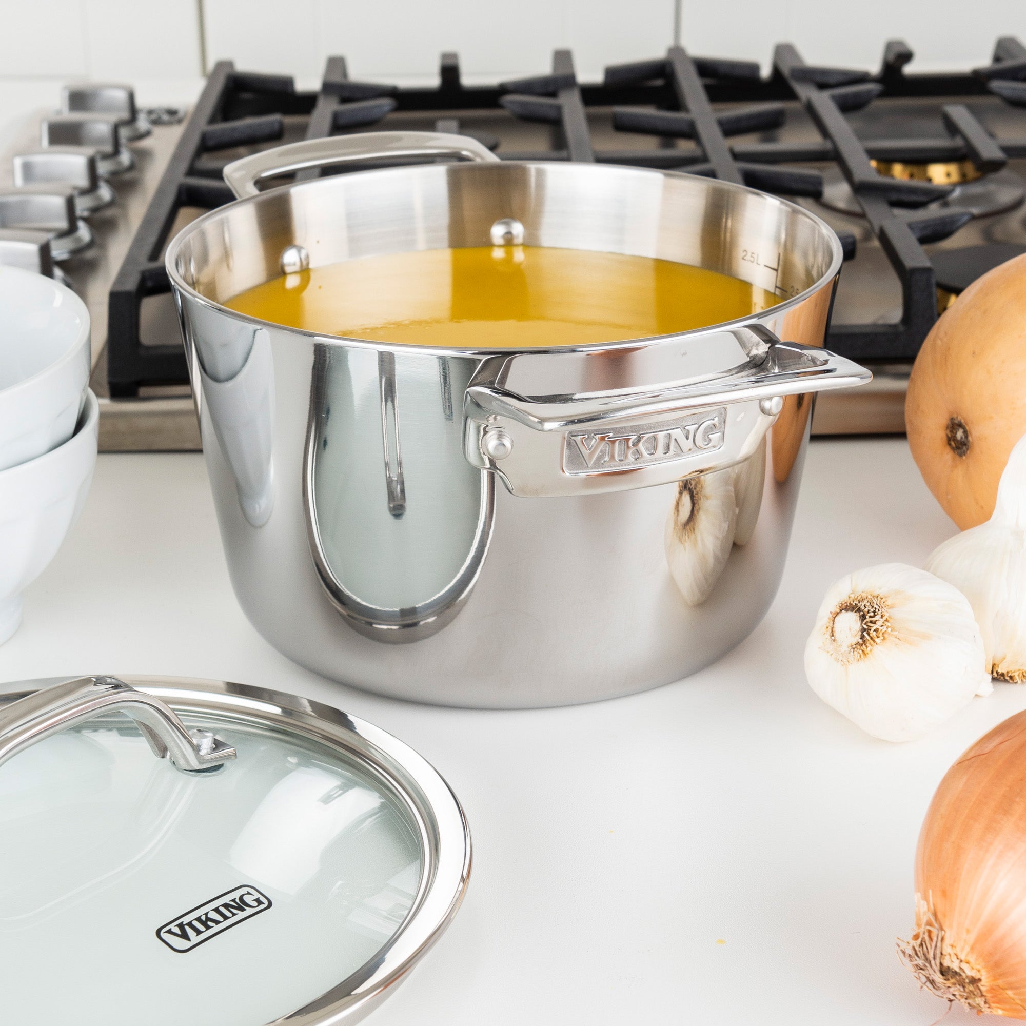 The Viking Contemporary 3-Ply Stainless Steel 3.4-Quart Soup Pot by Viking Culinary Products sits on a white countertop near a stove, filled with yellow soup and surrounded by onions, garlic, stacked bowls, and its glass lid.