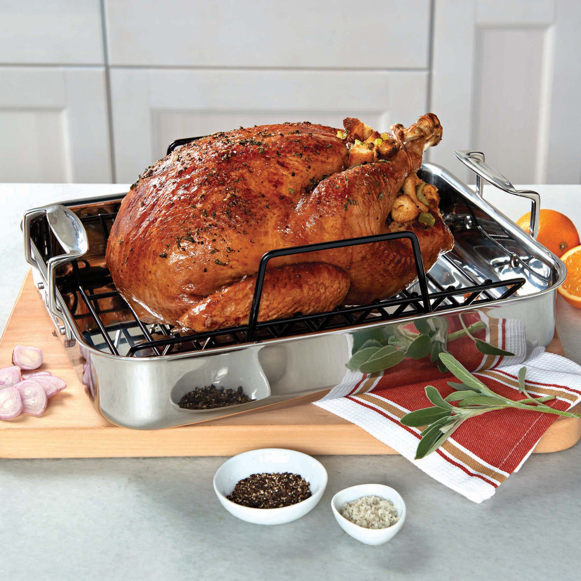 A roasted turkey rests in the Viking 3-Ply Stainless Steel Roaster with Rack by Viking Culinary Products, surrounded by herbs, shallots, pepper, and salt on a wooden board with a red-striped towel. The kitchen setting is bright and clean.
