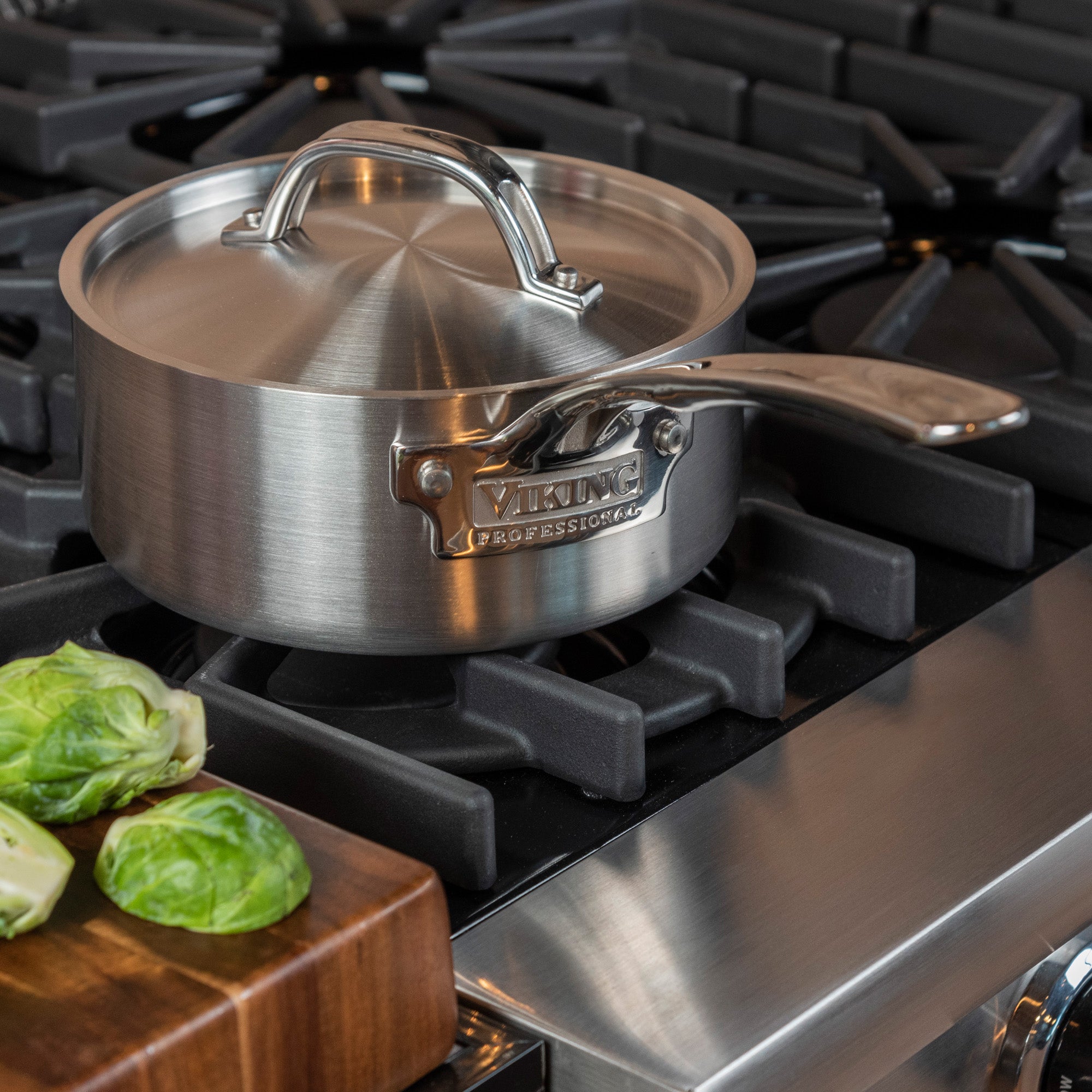 A Viking Culinary Products Professional 5-Ply 2-Quart Sauce Pan with Metal Lid, featuring stay-cool handles, sits on a gas stove. In the foreground, brussels sprouts are placed on a wooden cutting board.