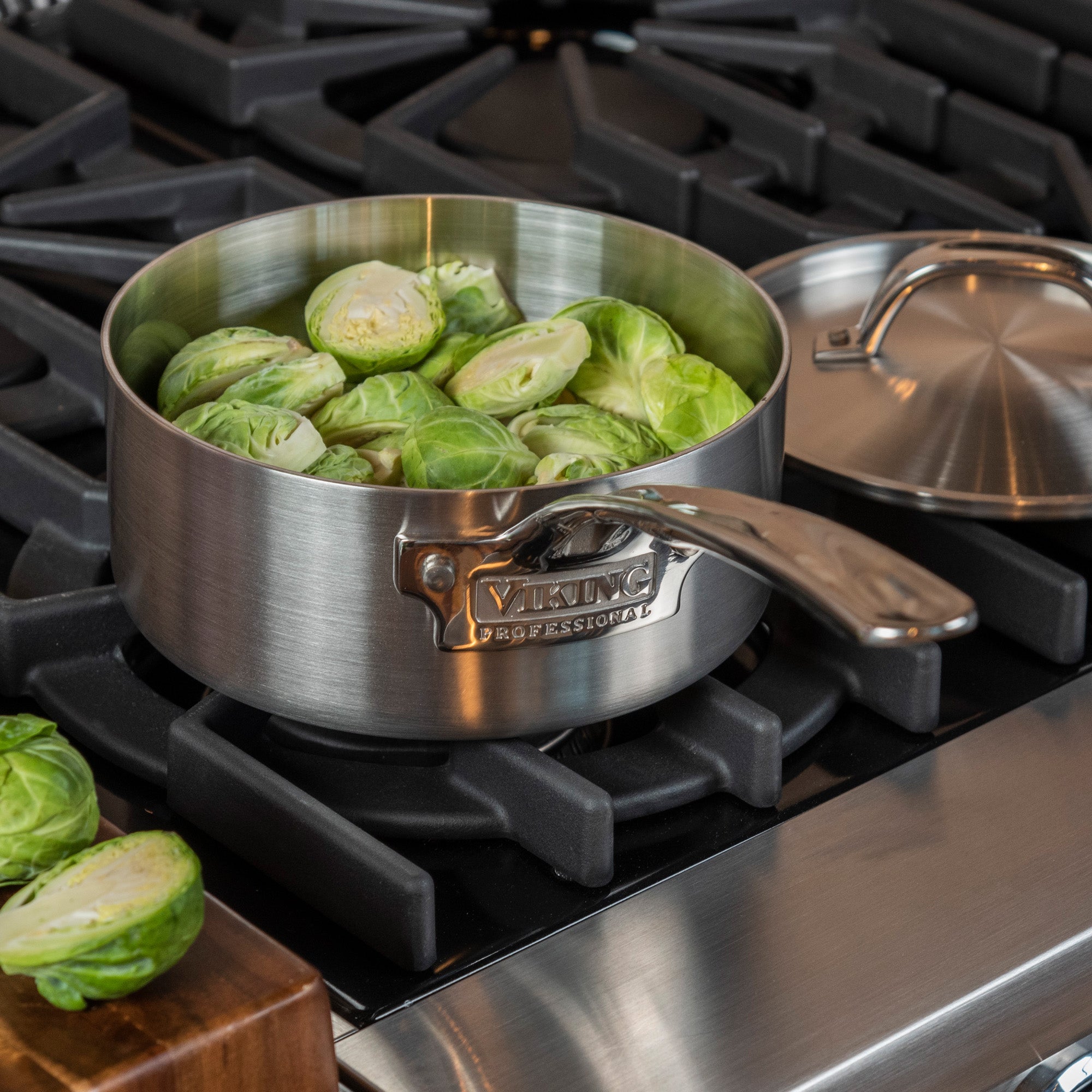 The Viking Professional 5-Ply 2-Quart Sauce Pan with Metal Lid from Viking Culinary Products holds Brussels sprouts on the stove, its lid set aside. Burner grates and a few extra Brussels sprouts rest on the wooden surface nearby.