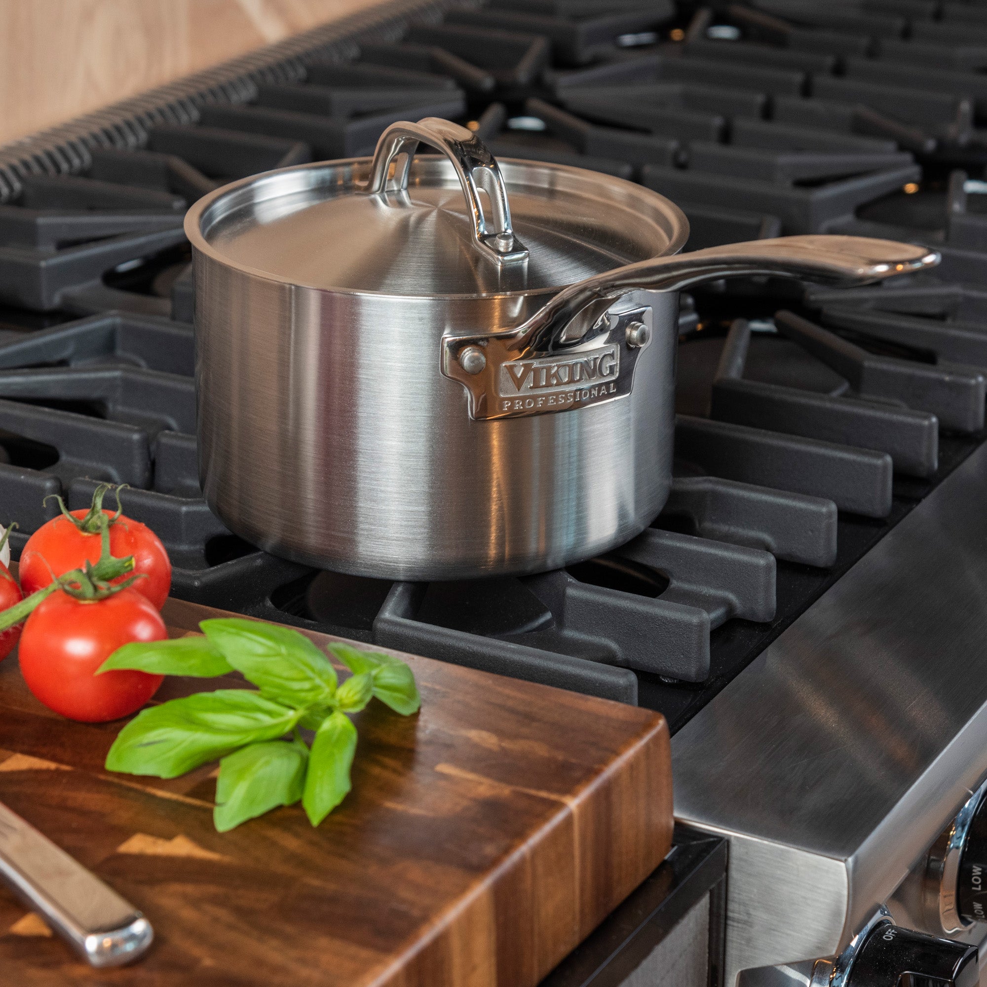The Viking Professional 5-Ply 3-Quart Sauce Pan with Metal Lid by Viking Culinary Products sits on a gas stove with black grates; a nearby wooden cutting board holds fresh tomatoes and basil. This saucepan is also dishwasher safe for easy cleanup.