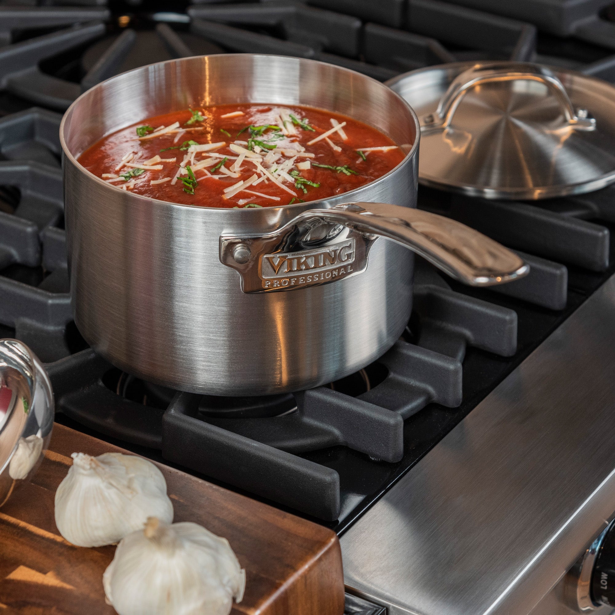 The Viking Culinary Products Professional 5-Ply 3-Quart Sauce Pan with Metal Lid simmers tomato soup, cheese, and herbs on a gas stove. The dishwasher-safe lid rests nearby, and two garlic bulbs sit on a cutting board beside the stove.