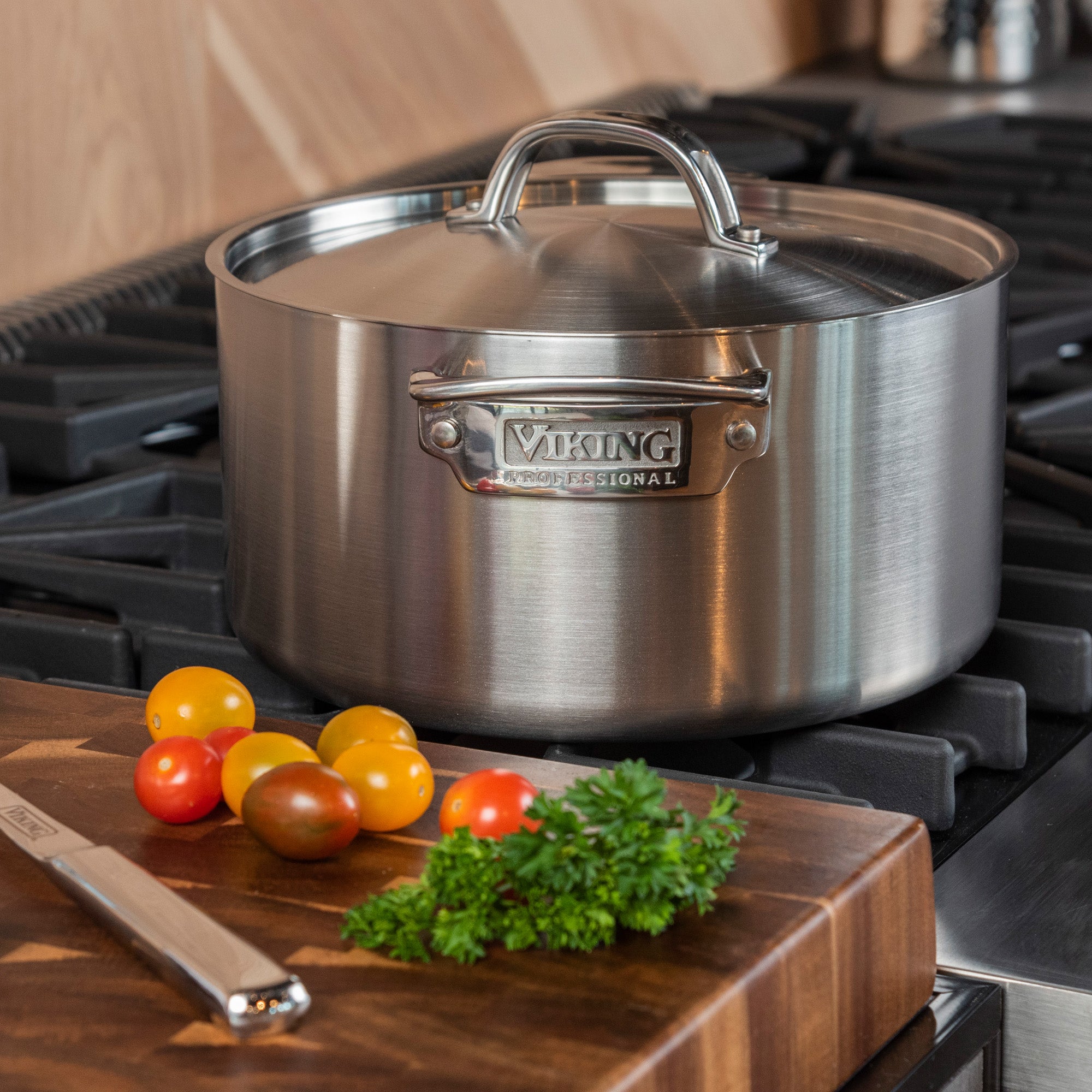 The Viking Professional 5-Ply Stainless Steel 6-Quart Stock Pot by Viking Culinary Products sits on a stove, lid on. In front, cherry tomatoes, parsley, and a knife are on a cutting board. This durable pot is dishwasher safe for easy cleanup.