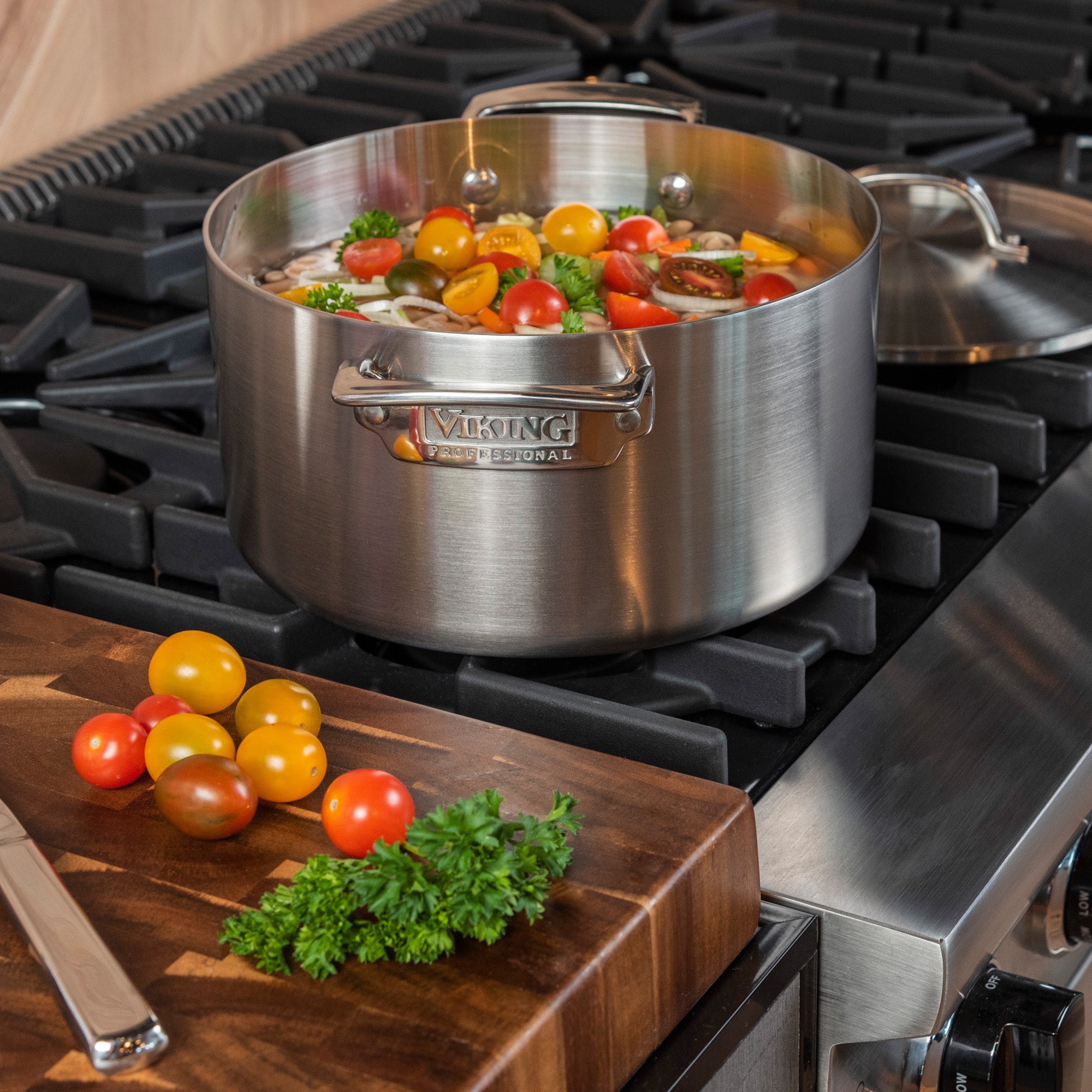 The Viking Professional 5-Ply Stainless Steel 6-Quart Stock Pot by Viking Culinary Products simmers tomatoes and herbs on a gas stove, while a nearby wooden cutting board holds tomatoes, parsley, and a knife. Dishwasher safe for easy cleanup.