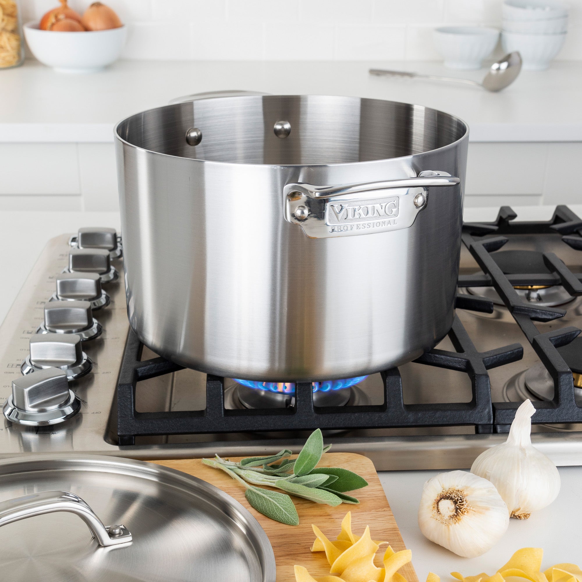 The Viking Professional 5-Ply Stainless Steel 8-Quart Stock Pot by Viking Culinary Products sits on a gas stove with a blue flame, surrounded by garlic, sage, uncooked pasta, and its lid. Eggs, bowls, and utensils are seen on the kitchen counter in the background.