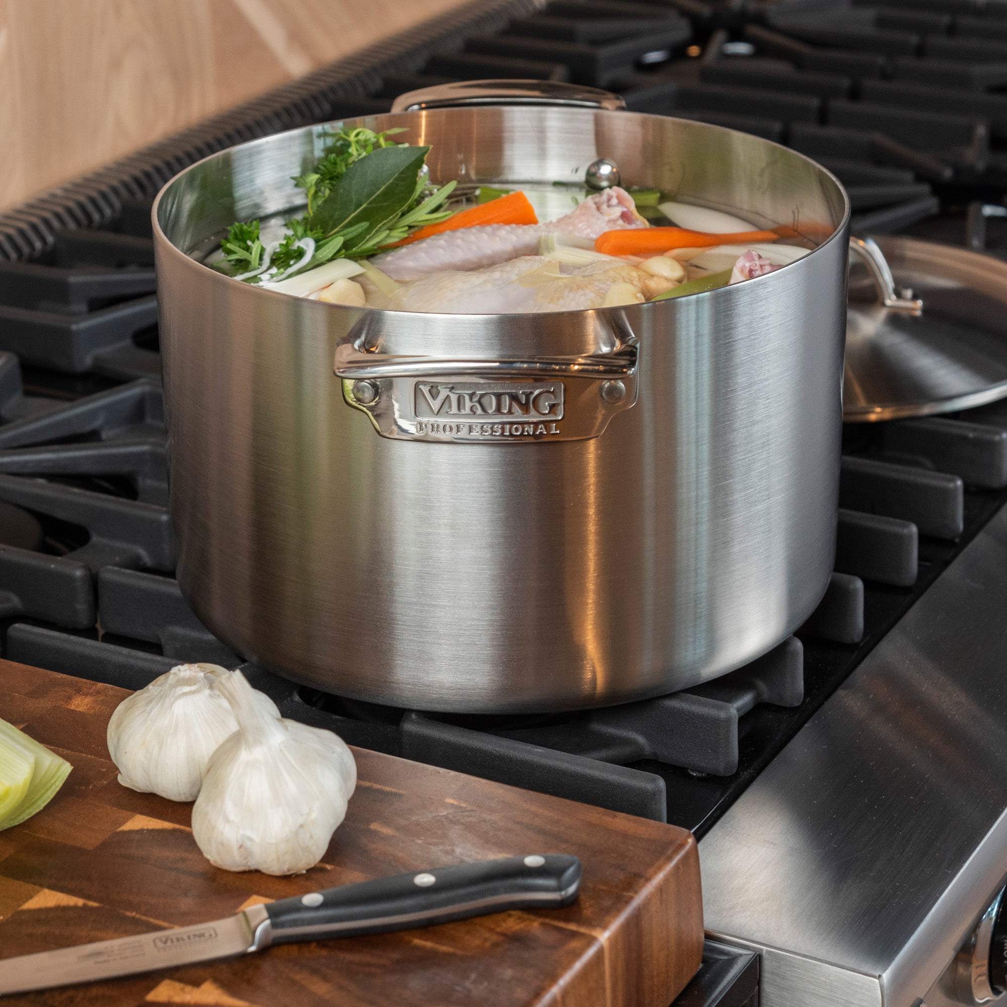 A Viking Culinary Products Professional 5-Ply Stainless Steel 8-Quart Stock Pot sits on the stove with vegetables, herbs, and chicken. On a wooden cutting board nearby are two garlic heads, a knife, and some celery.