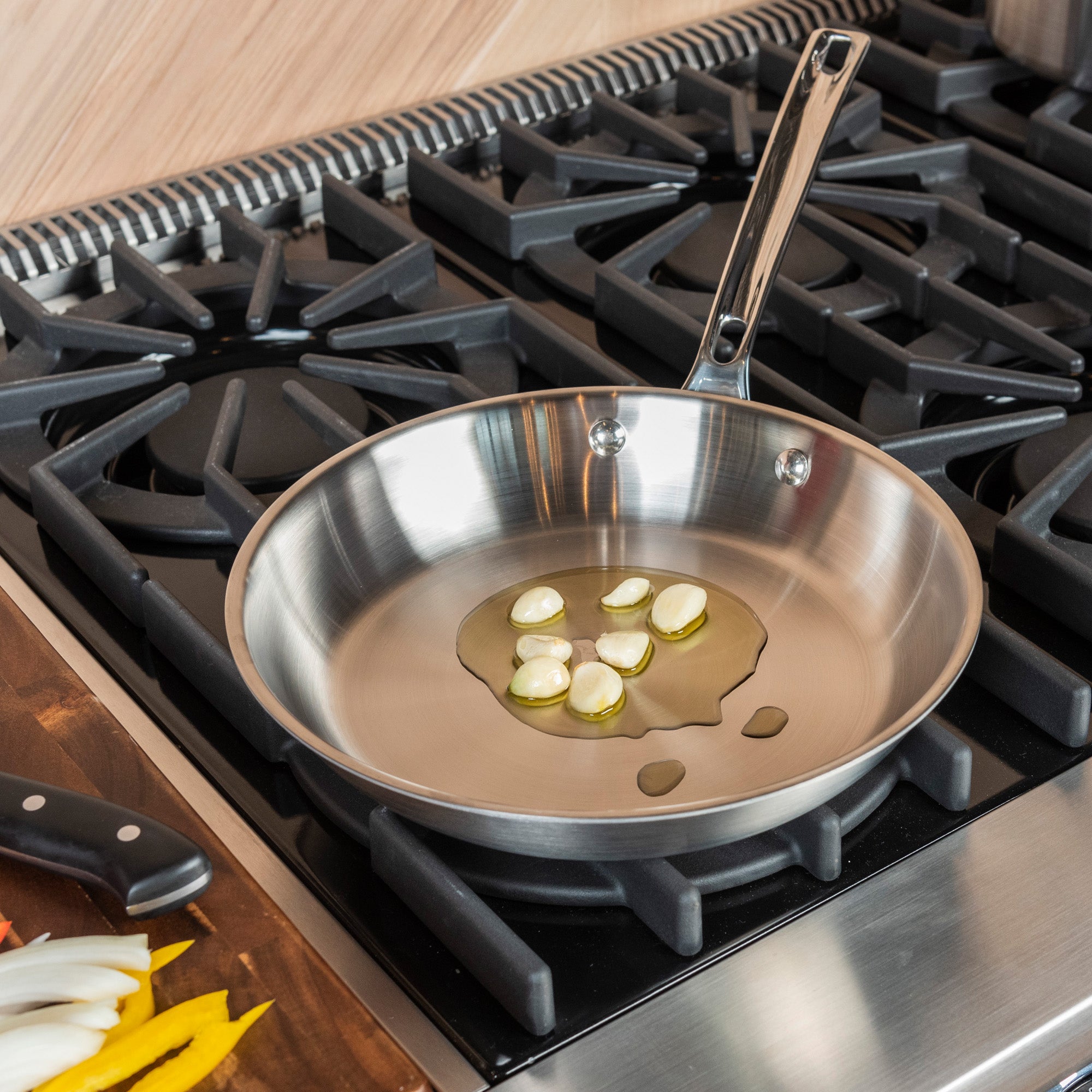 The Viking Culinary Products Professional 5-Ply 5-Piece Starter Cookware Set features a stainless steel frying pan heating garlic and oil on a gas stovetop, with a knife and sliced vegetables nearby—showcasing premium cookware in action.