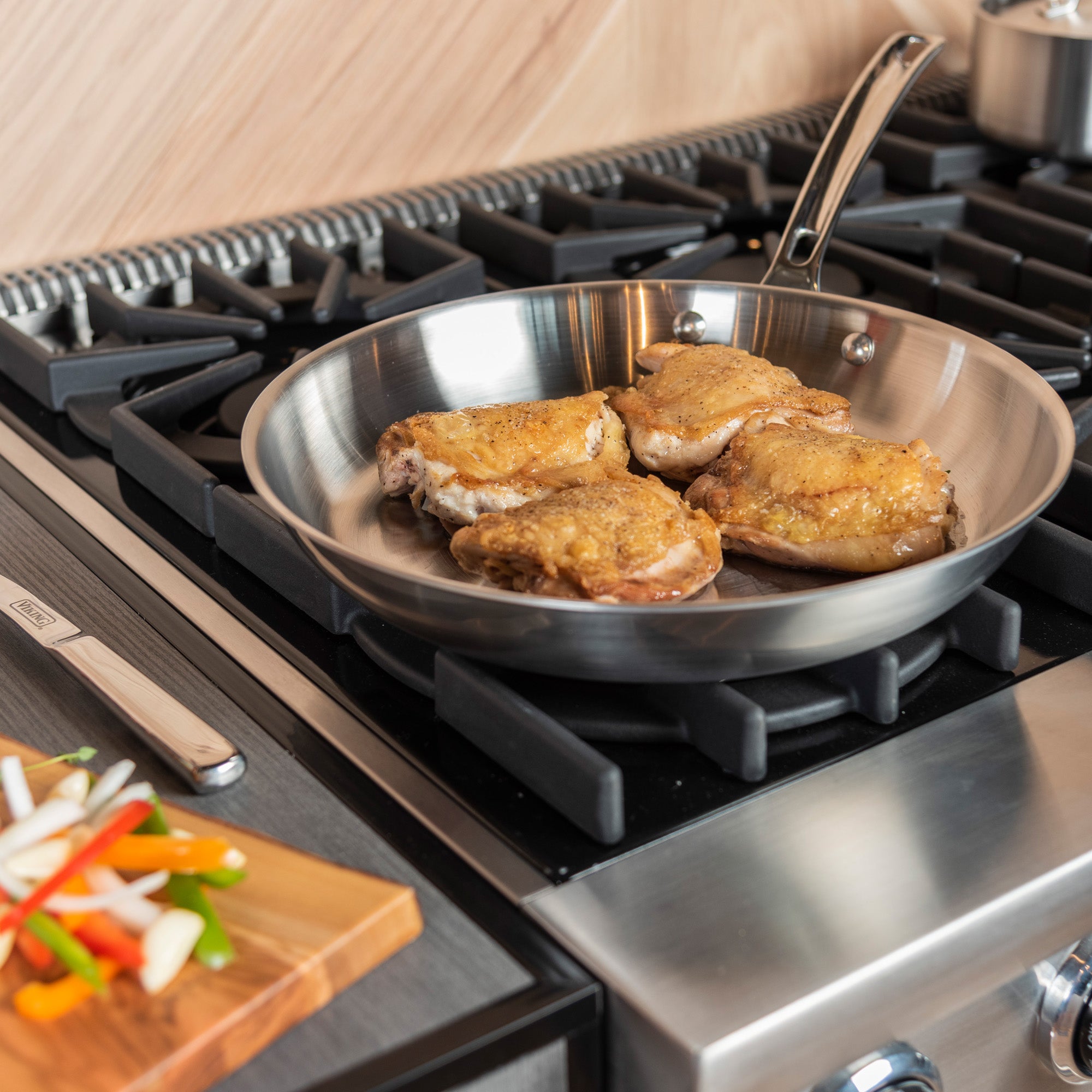 Chicken thighs sizzle in the Viking Professional 5-Ply 12-Inch Fry Pan by Viking Culinary Products on a gas stove, while a wooden cutting board with sliced bell peppers and onions sits nearby in the modern kitchen.