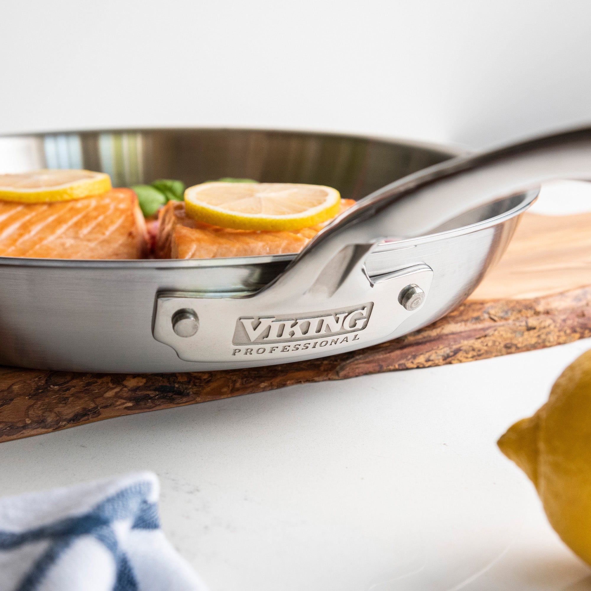 A Viking Culinary Products Professional 5-Ply 12-Inch Fry Pan with Stay-Cool Handle holds salmon fillets topped with lemon slices on a wooden board, with a cut lemon and towel visible in the foreground.