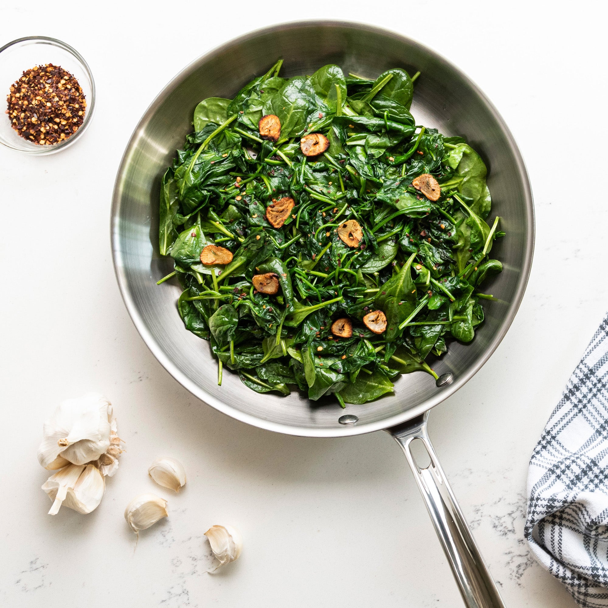 The Viking Professional 5-Ply 12-Inch Fry Pan by Viking Culinary Products, featuring Stay-Cool Handles, holds sautéed spinach and sliced garlic on a white surface beside red pepper flakes, garlic cloves, and a blue-and-white kitchen towel.