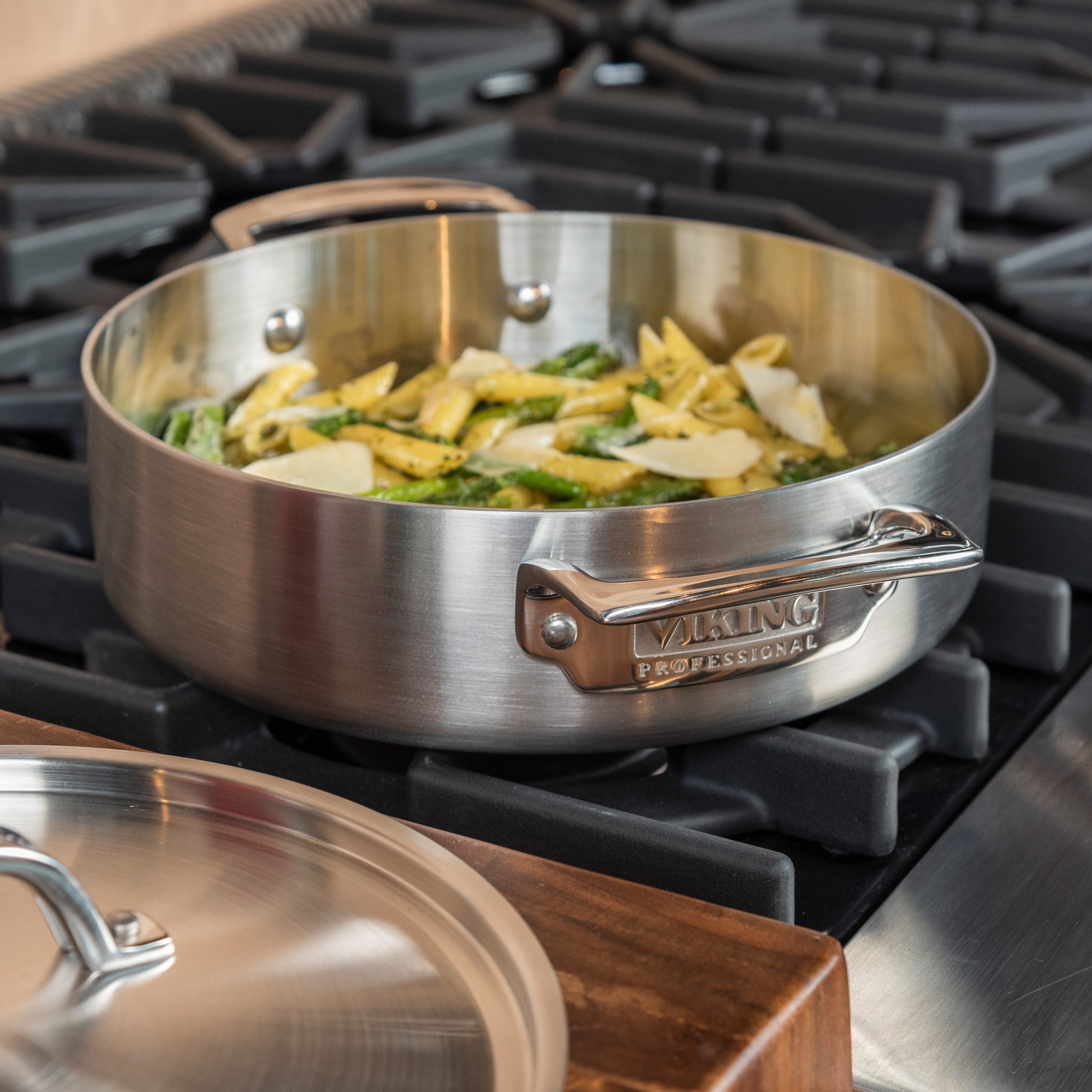 A Viking Culinary Products Professional 5-Ply Stainless Steel 3.4-Quart Casserole Pan with sautéed vegetables sits on a gas stove, while its matching, dishwasher-safe lid rests on the counter nearby.