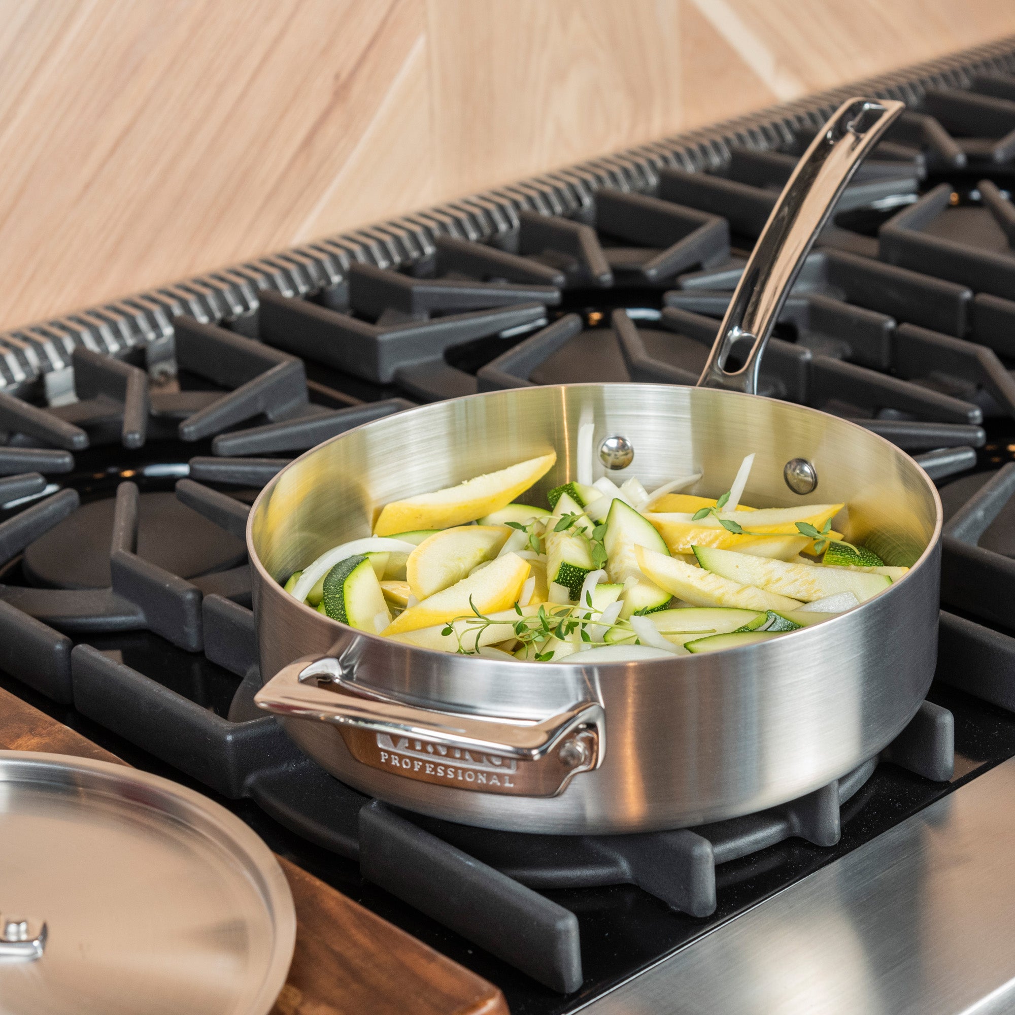The Viking Professional 5-Ply Stainless Steel 3.4-Quart Sauté Pan by Viking Culinary Products holds sliced zucchini and onions on a gas stove, with its metal lid resting on the counter nearby.