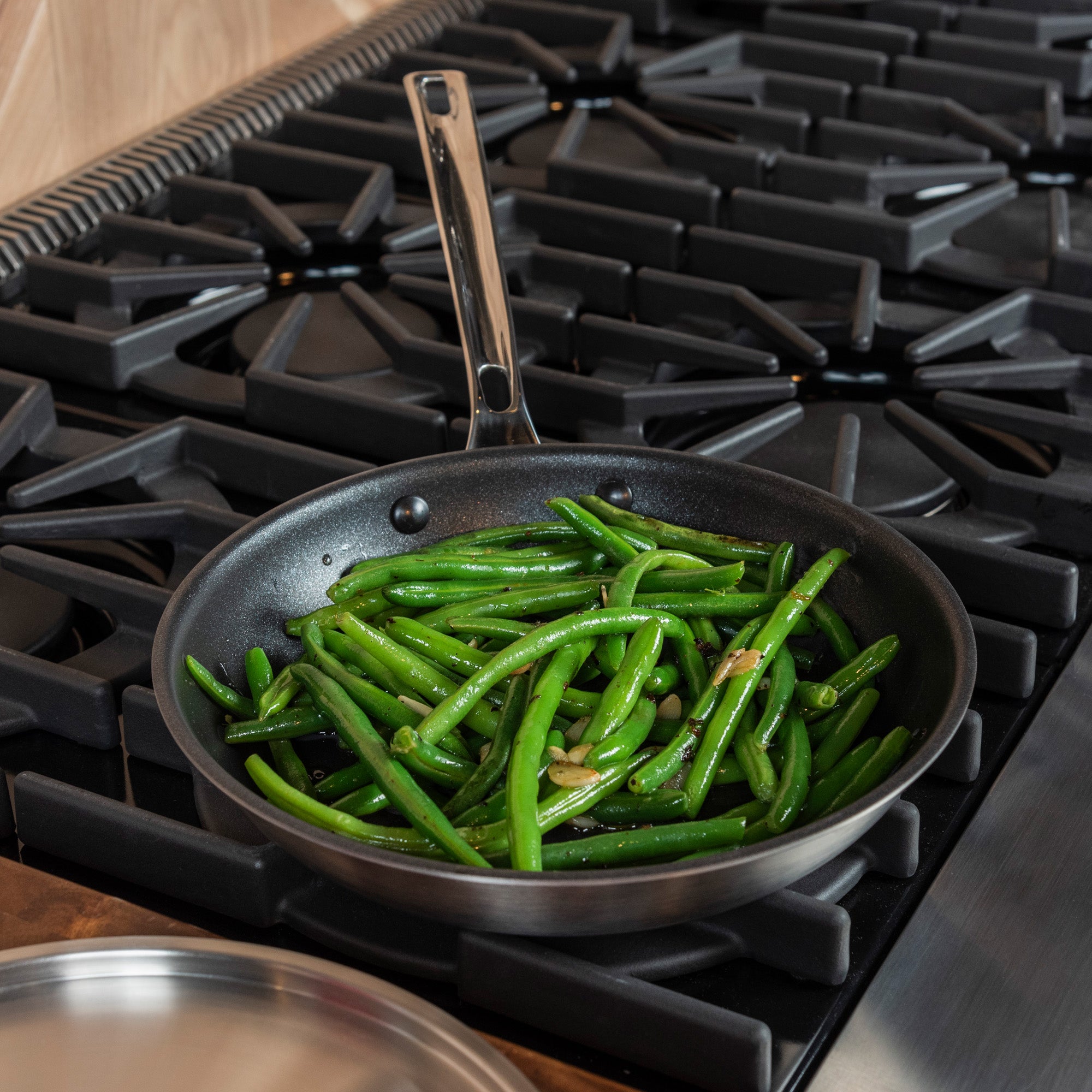 The Viking Professional 5-Ply Stainless Steel Covered Nonstick 10-Inch Fry Pan by Viking Culinary Products, filled with sautéed green beans and garlic, rests on a black gas stove beside its stainless steel stay-cool lid.