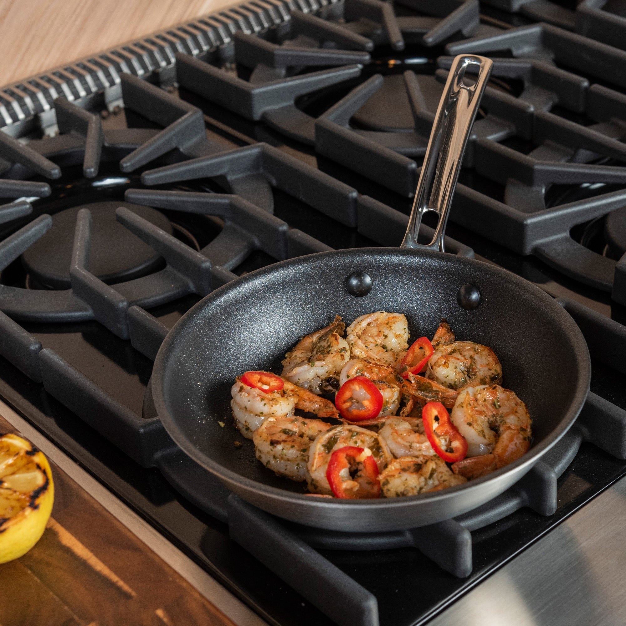 A Viking Professional 5-Ply 8-Inch Nonstick Fry Pan by Viking Culinary Products, with cooked shrimp and sliced red chili, sits on a gas stove. Thanks to its Stay-Cool Handle, cooking and serving are easy. A grilled lemon half rests on a wooden countertop nearby.