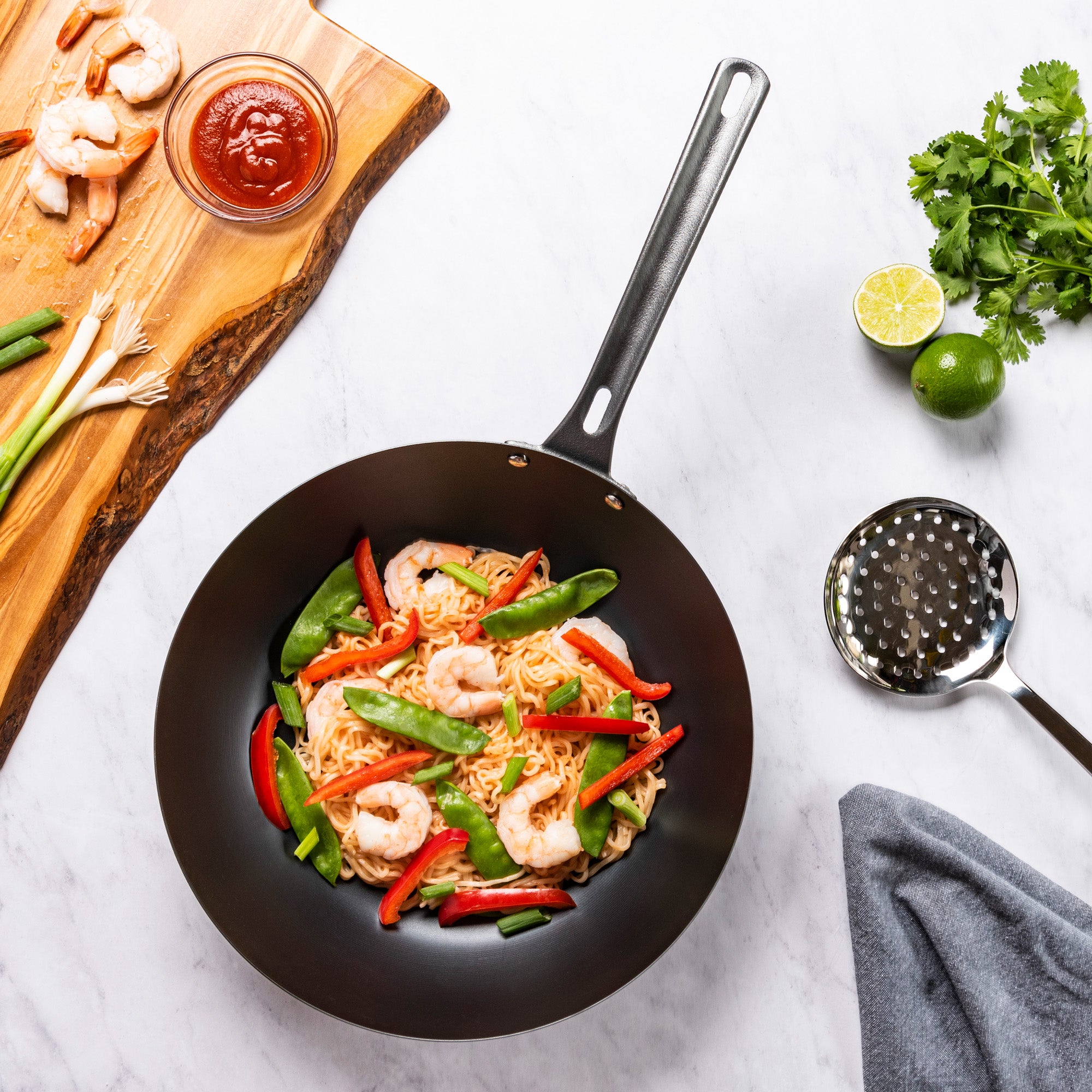 A Viking Culinary Products Viking Blue Carbon Steel 12-Inch Wok filled with shrimp, noodles, snap peas, and red bell peppers sits on a white surface next to a cutting board with shrimp and green onions, a bowl of sauce, cilantro, and lime.