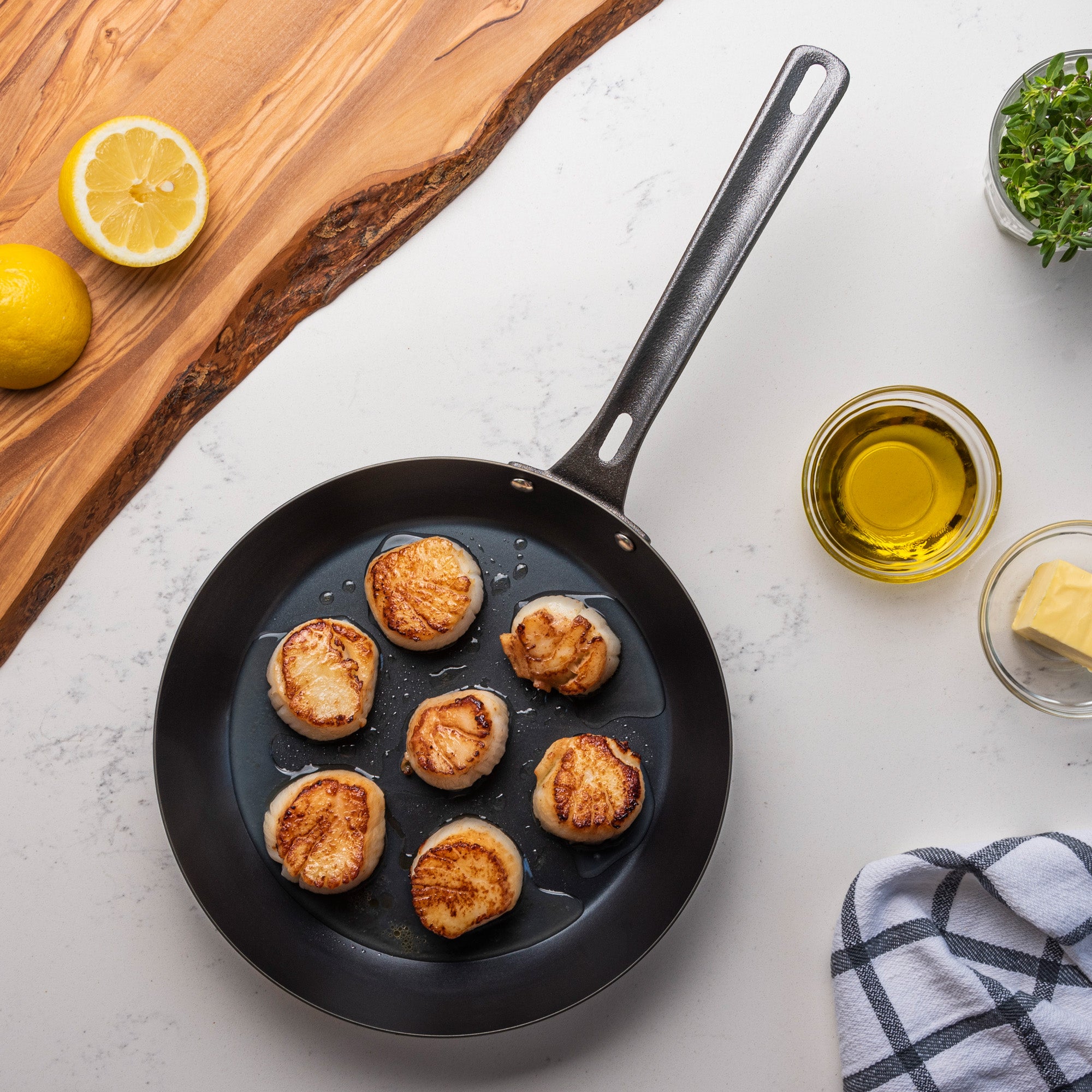 A Viking Blue Carbon Steel 10-Inch Fry Pan by Viking Culinary Products, pre-seasoned and black, holds seven seared scallops on a white countertop beside olive oil, butter, half a lemon, green herbs, and a checkered cloth.