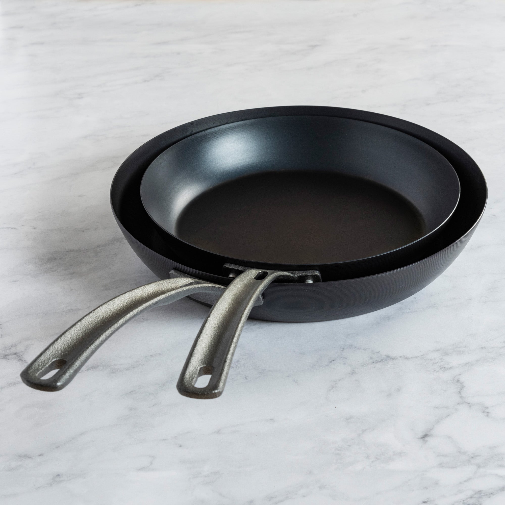 Two Viking Culinary Products blue carbon steel fry pans from the Viking 2-Piece Set are stacked on a light marble countertop, featuring metal handles.