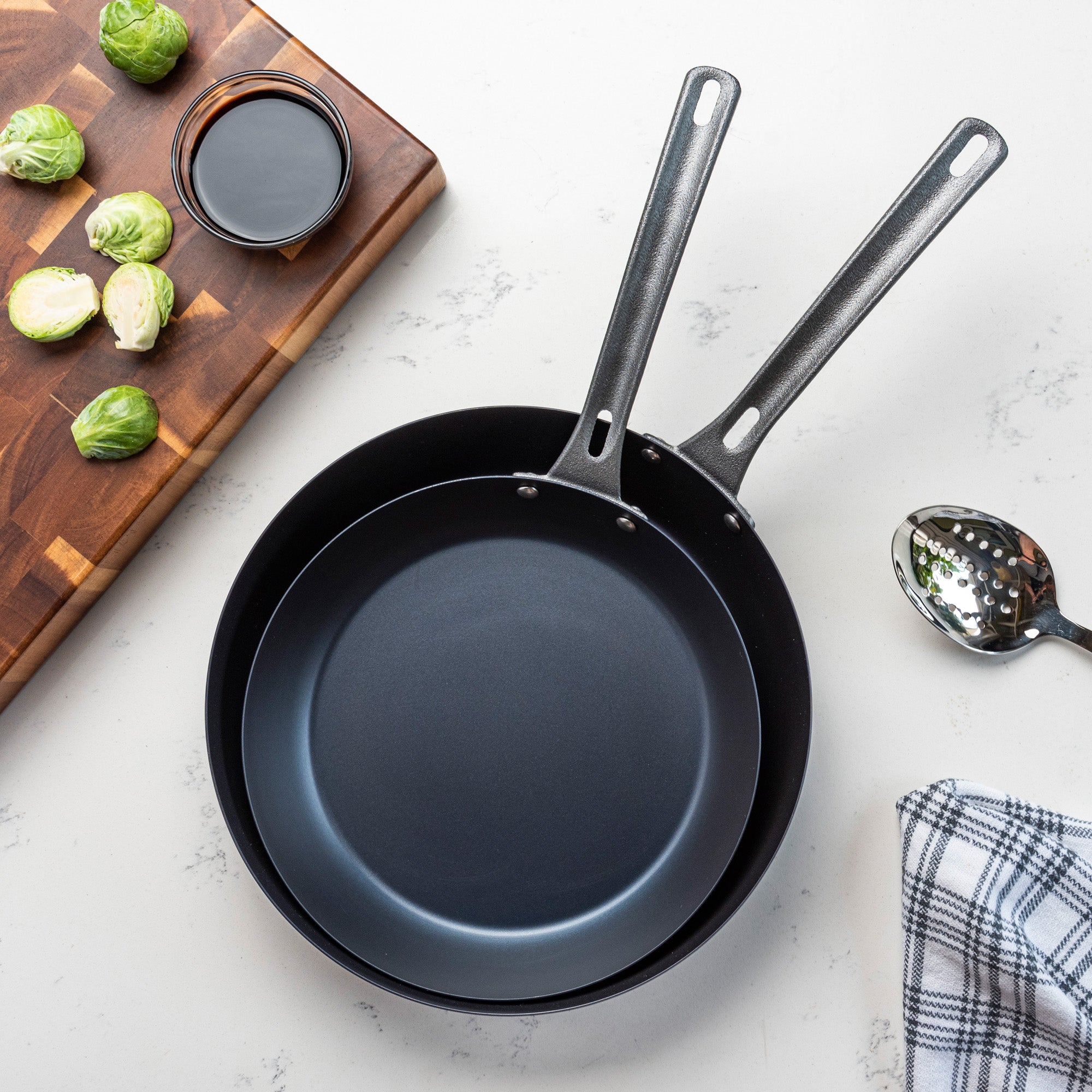 The Viking 2-Piece Blue Carbon Steel Fry Pan Set by Viking Culinary Products sits on a light countertop beside a slotted spoon, plaid towel, cutting board with Brussels sprouts, and a bowl of dark sauce—ideal for any kitchen.