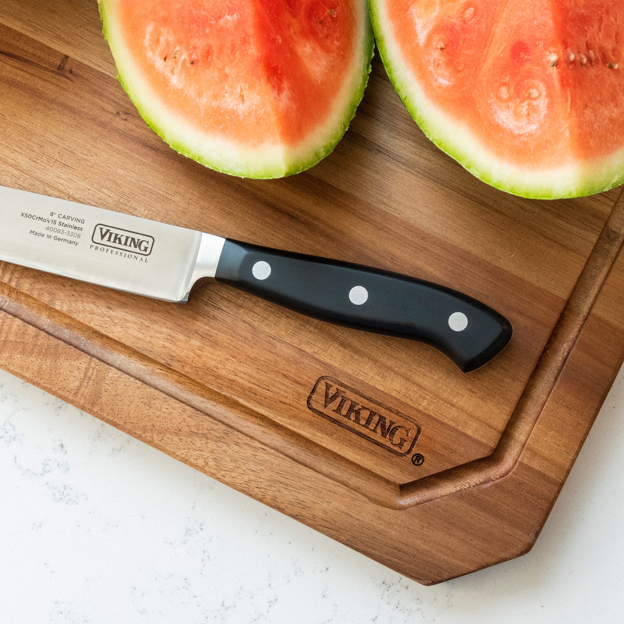 A Viking brand knife and half a watermelon sit on the Viking Acacia Carving Board with Juice Groove by Viking Culinary Products, displayed on a white countertop.