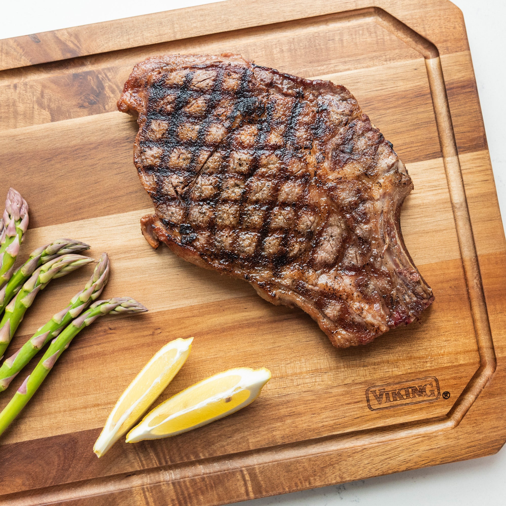 A grilled steak sits on the Viking Acacia Carving Board with Juice Groove by Viking Culinary Products, garnished with three asparagus spears and two lemon wedges.
