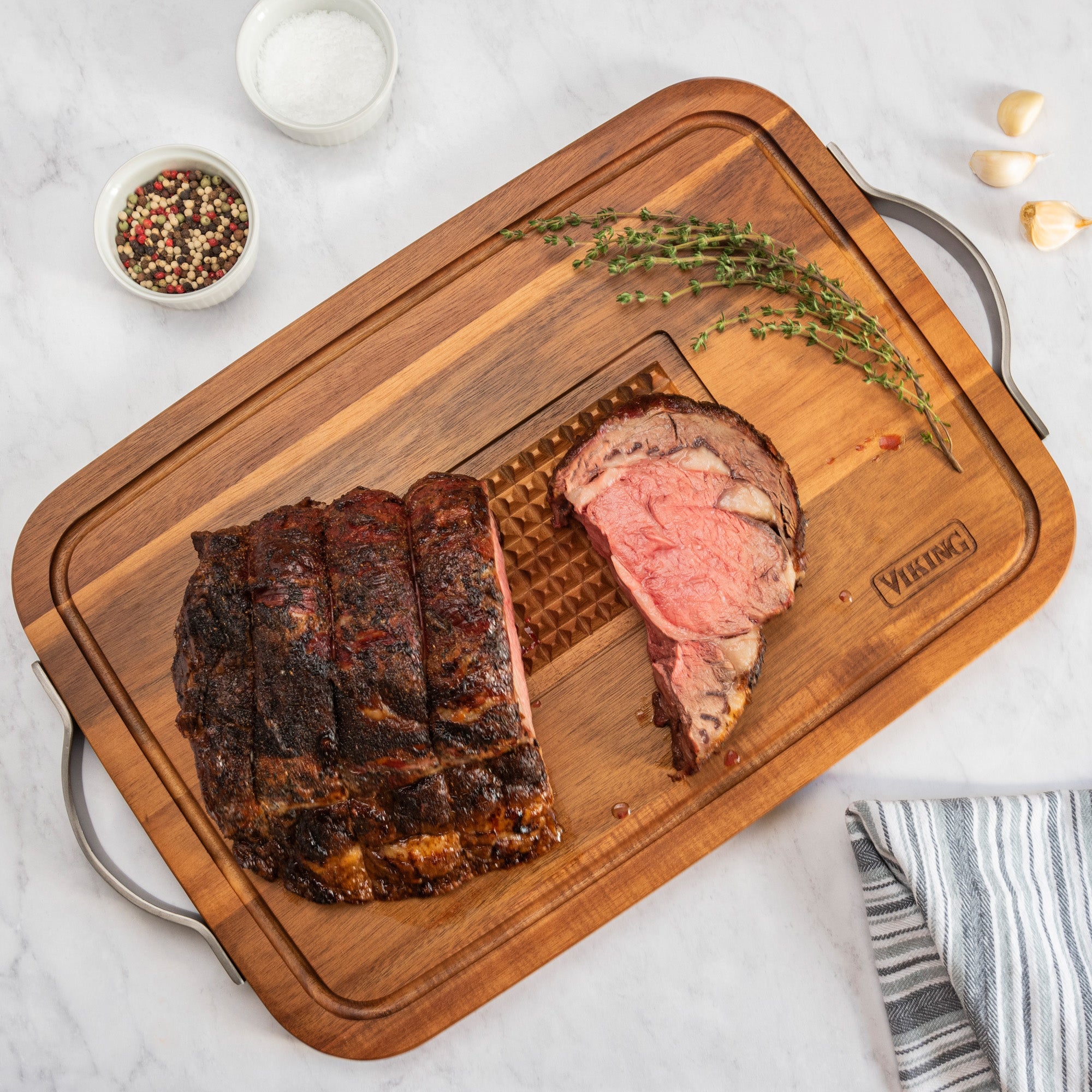 A sliced roasted beef ribeye sits on the Viking Acacia Carving Board with Juice Well and Metal Handle by Viking Culinary Products, surrounded by thyme, garlic cloves, and bowls of salt and peppercorns for a stylish, functional presentation.