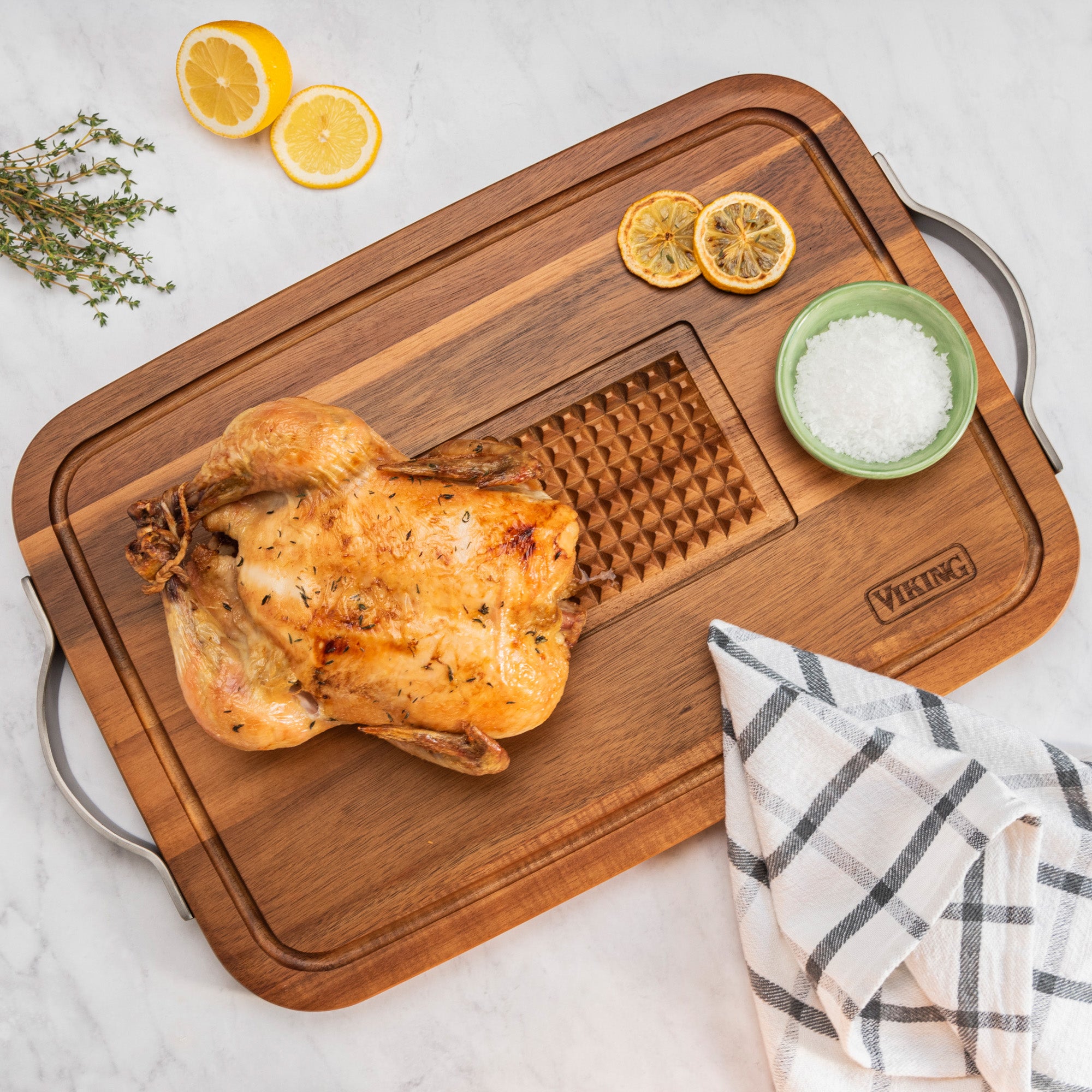 A roasted chicken rests on the Viking Acacia Carving Board with Juice Well and Metal Handle by Viking Culinary Products, surrounded by a towel, salt in a green bowl, fresh thyme, and lemon slices on a marble surface.