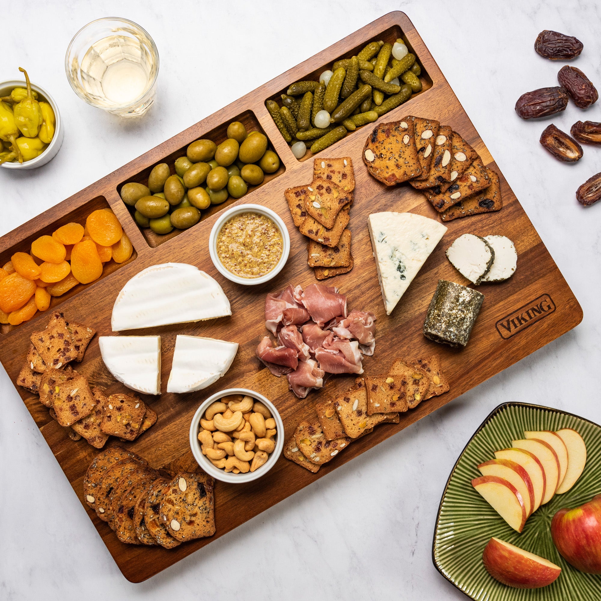 A Viking Acacia 20-Inch Reversible Butcher Block by Viking Culinary Products holds cheeses, salami, crackers, green olives, pickles, dried apricots, nuts, mustard, and white wine beside a plate of sliced apples and dates.