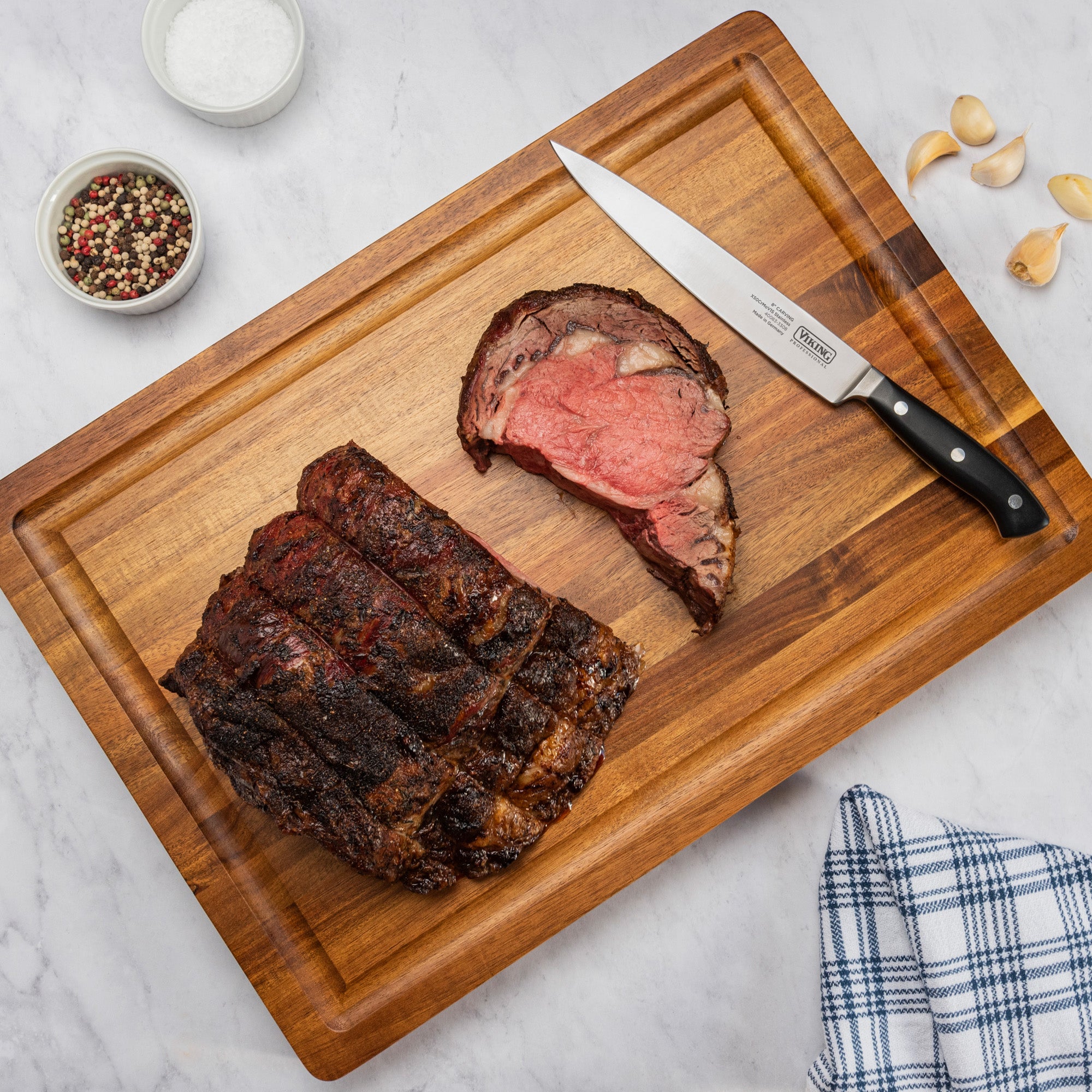 Sliced roast beef sits on the Viking Culinary Products Acacia 20-Inch Reversible Butcher Block Prep and Carving Board, with a carving knife, whole peppercorns, salt, garlic cloves, and a blue-and-white towel on marble.