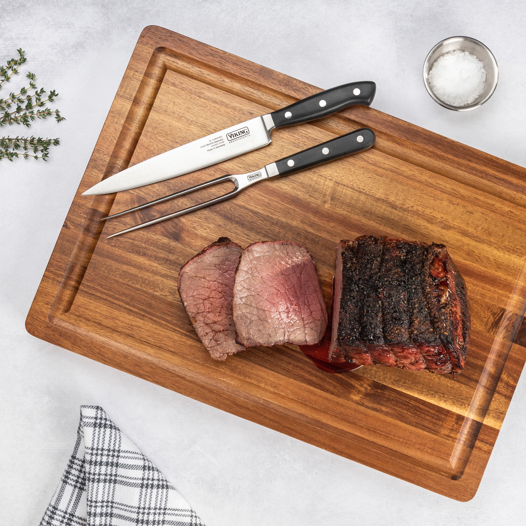 Sliced roast beef on a wooden cutting board with the Viking Professional 2-Piece Carving Set by Viking Culinary Products, plus a bowl of salt, fresh herbs, and a checkered kitchen towel on a light surface.