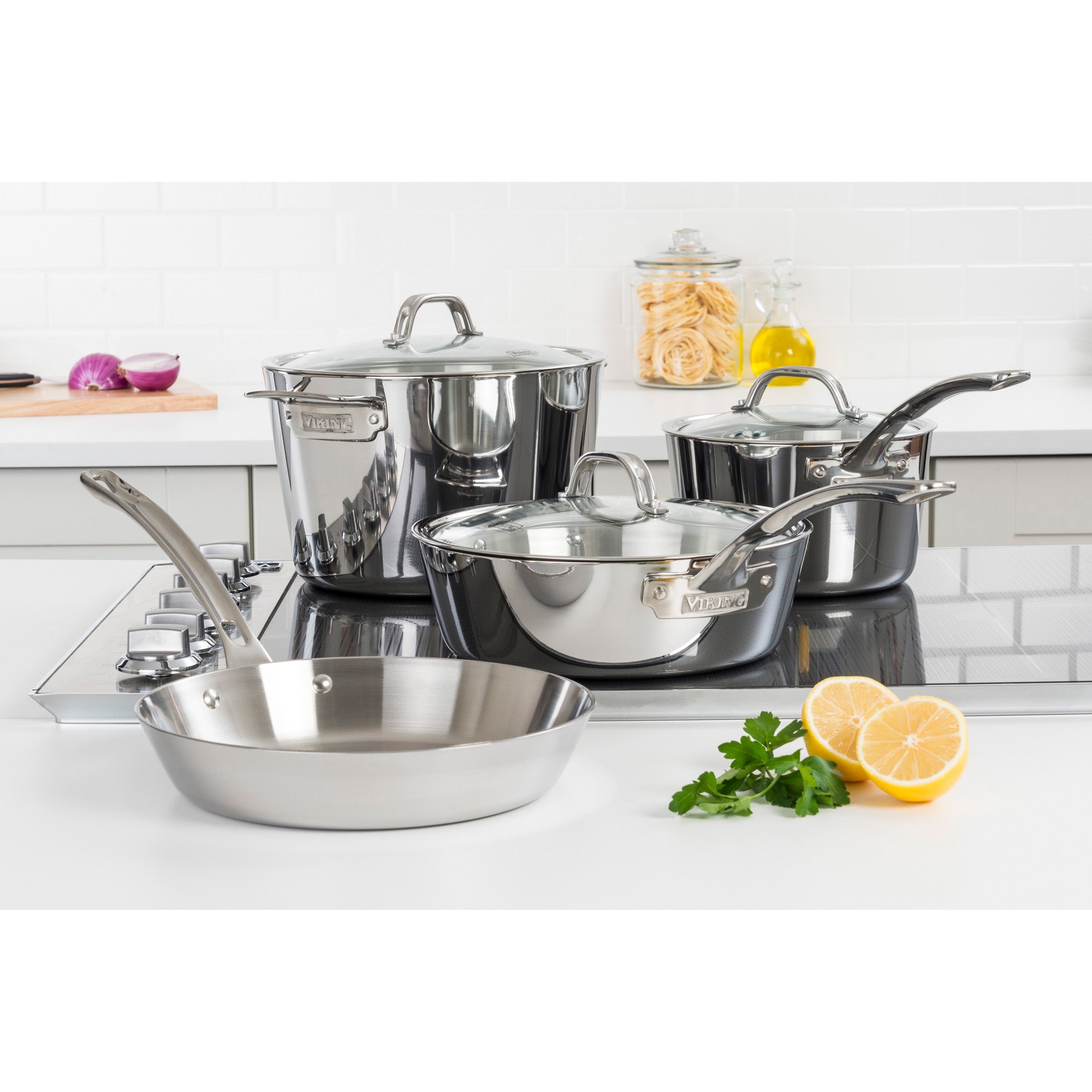 The Viking Contemporary 3-Ply Stainless Steel 7-Piece Cookware Set with Glass Lids by Viking Culinary Products is arranged on a modern kitchen stovetop, with fresh parsley, a halved lemon, olive oil, and jarred pasta nearby. Induction compatible and stylish.