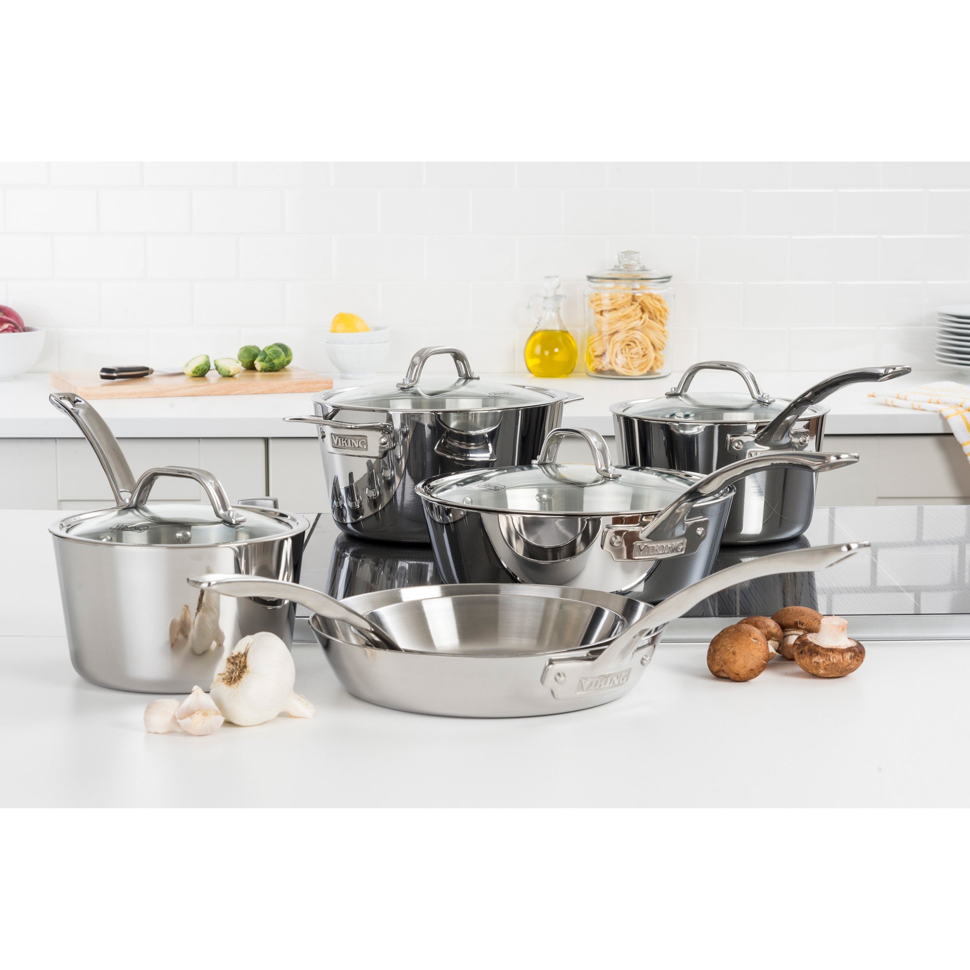 The Viking Contemporary 3-Ply Stainless Steel 10-Piece Cookware Set with Glass Lids by Viking Culinary Products is displayed on a kitchen counter, surrounded by garlic, mushrooms, limes, pasta in a glass jar, and oil bottles in the background.
