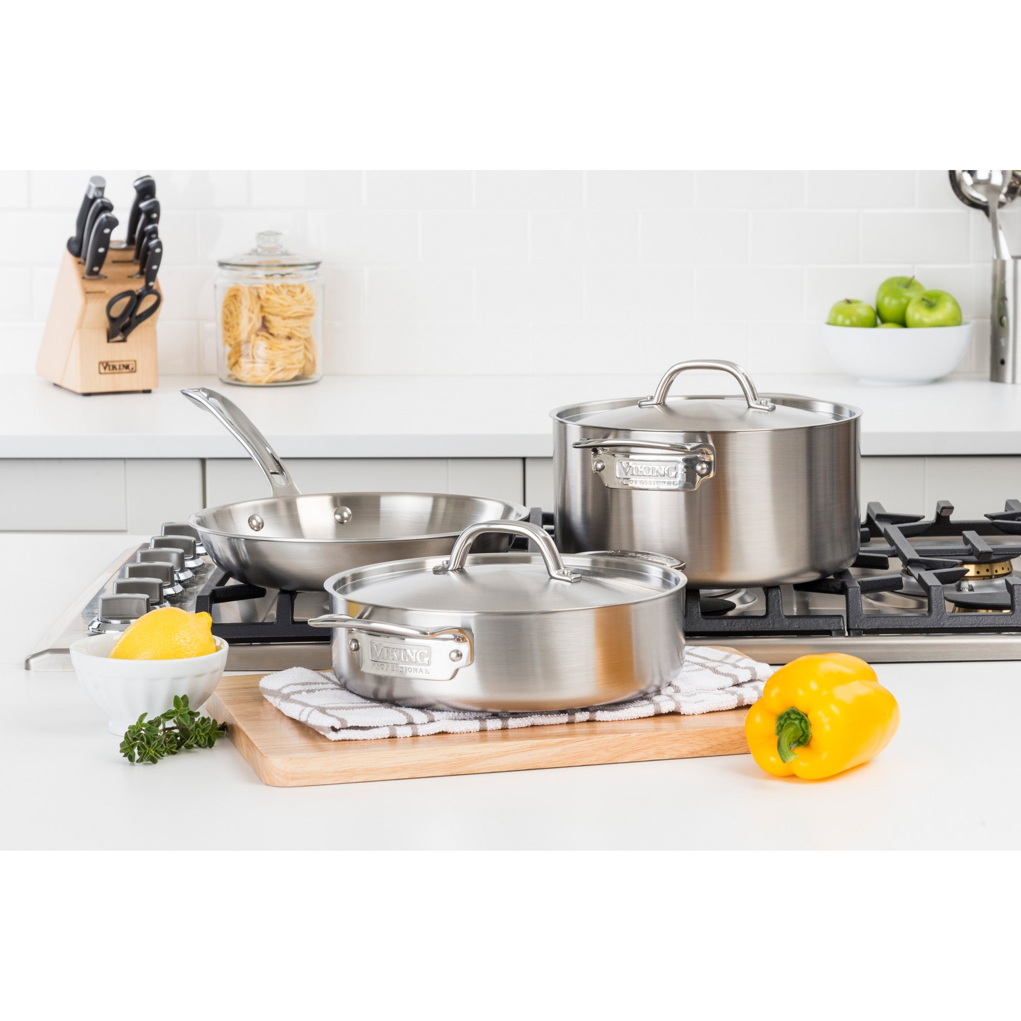 The Viking Culinary Products Professional 5-Ply 5-Piece Starter Cookware Set with Stay-Cool Handles is displayed on a stovetop and cutting board in a bright kitchen, surrounded by a yellow bell pepper, lemons with herbs, and a knife block.