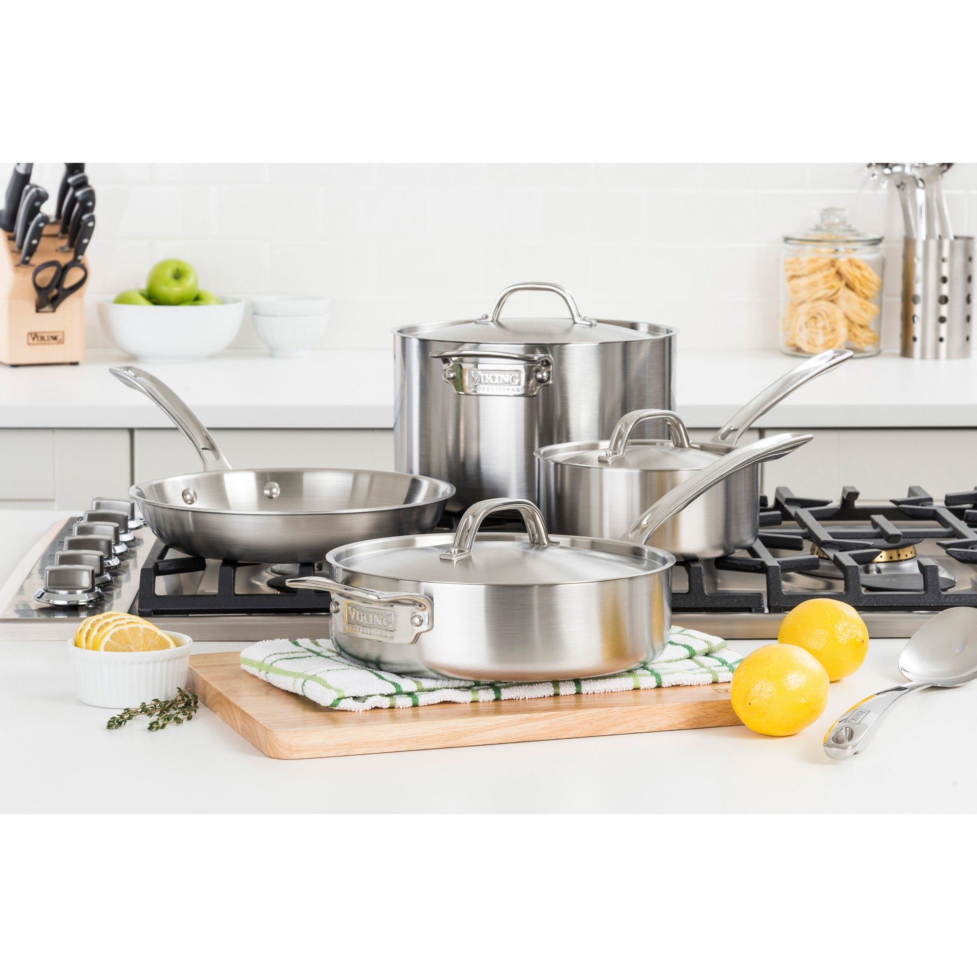The Viking Professional 5-Ply 7-piece Cookware Set by Viking Culinary Products, featuring Stay-Cool Handles, is displayed on a modern kitchen stovetop and counter with lemons, herbs, a dish towel, utensils, and ingredients nearby.